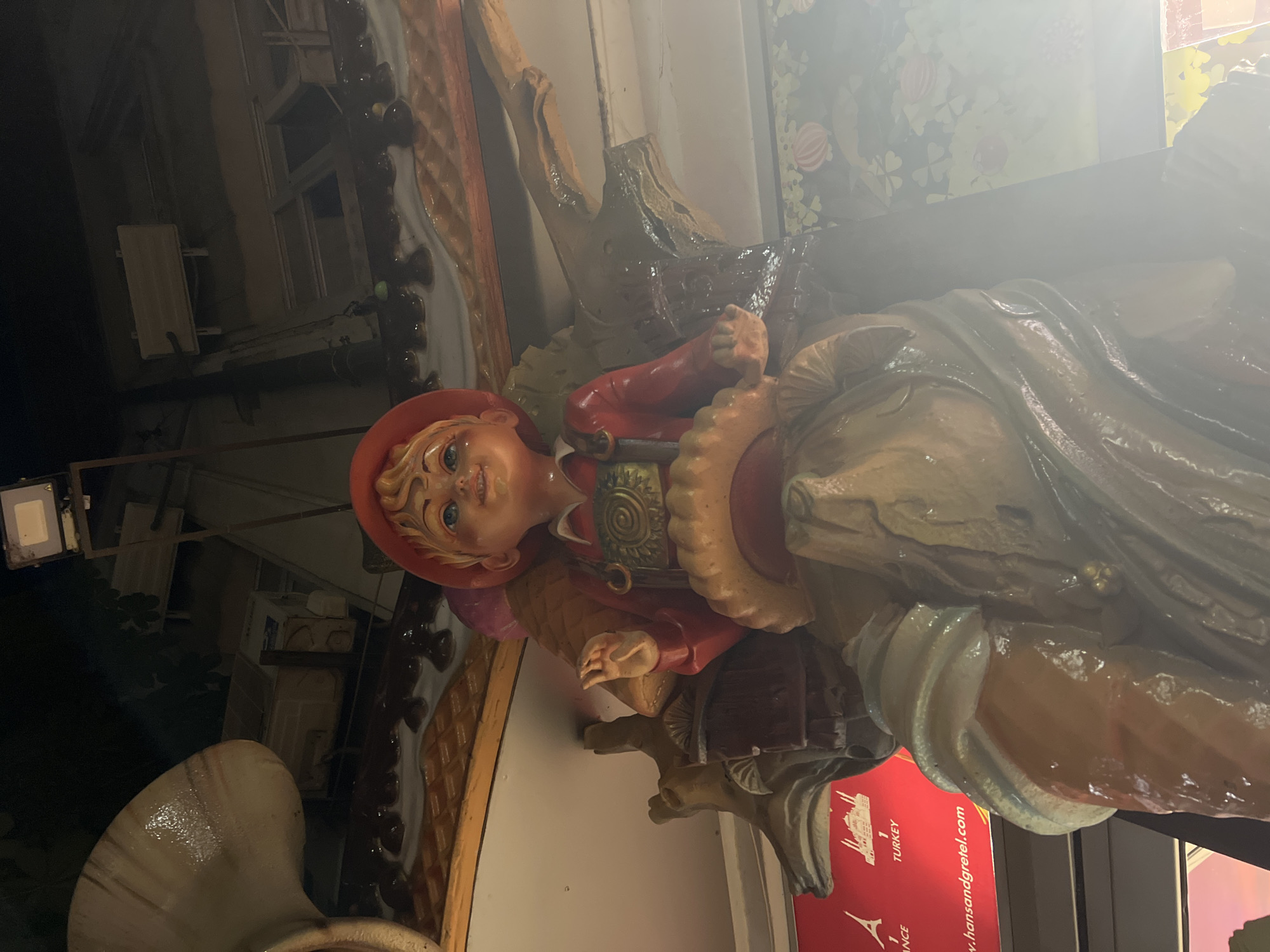 The image shows a close-up of a detailed, painted statue of a young boy wearing a red hat and traditional German attire. The boy has blue eyes, blond hair, and a cheerful expression. He is seated on what appears to be a wooden structure, possibly a carved animal or a tree trunk. The statue is part of a larger decorative display. In the background, there is a red sign with text that includes "TURKEY" and "FRANCE," and a website address that appears to be "www.hansandgreta.com". The lighting suggests the image was taken indoors, likely in a themed attraction or shop. The overall style of the statue is reminiscent of fairy tale characters. The presence of the text indicating "TURKEY" and "FRANCE" along with the website related to Hansel and Gretel suggests the context is a tourist attraction or themed environment. The statue itself is the central focus, with detailed rendering of clothing and facial features.
