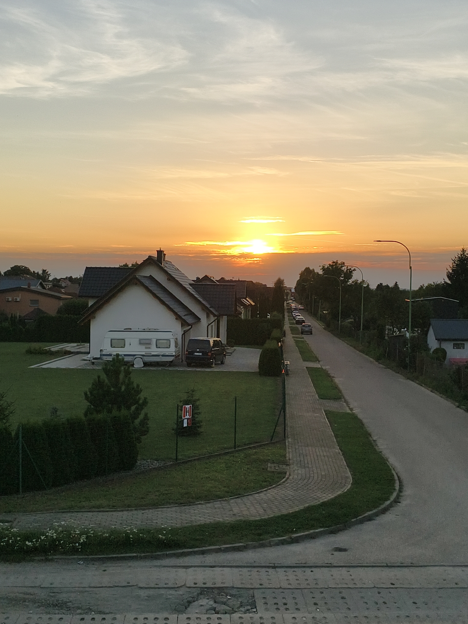 The image depicts a residential street at sunset in Tczew, Poland. The sky is filled with warm, orange and yellow hues, with wispy clouds. A single-family house with a white facade and dark tiled roof is prominent on the left side of the image. In front of the house, a white caravan is parked next to a black van. A neatly mowed lawn with a hedge borders the property.
The street itself is paved and curves to the right. Several cars are parked along the left side of the street. Streetlights with modern, curved designs are visible along the sidewalk. The sidewalk is made of brick pavers and is bordered by grass. Further down the street, more houses and trees are visible. A small red and white sign with an 'H' inside a circle is situated on a fence post in the foreground. The foreground also shows a concrete structure with circular perforations. The overall impression is a peaceful, suburban evening scene.