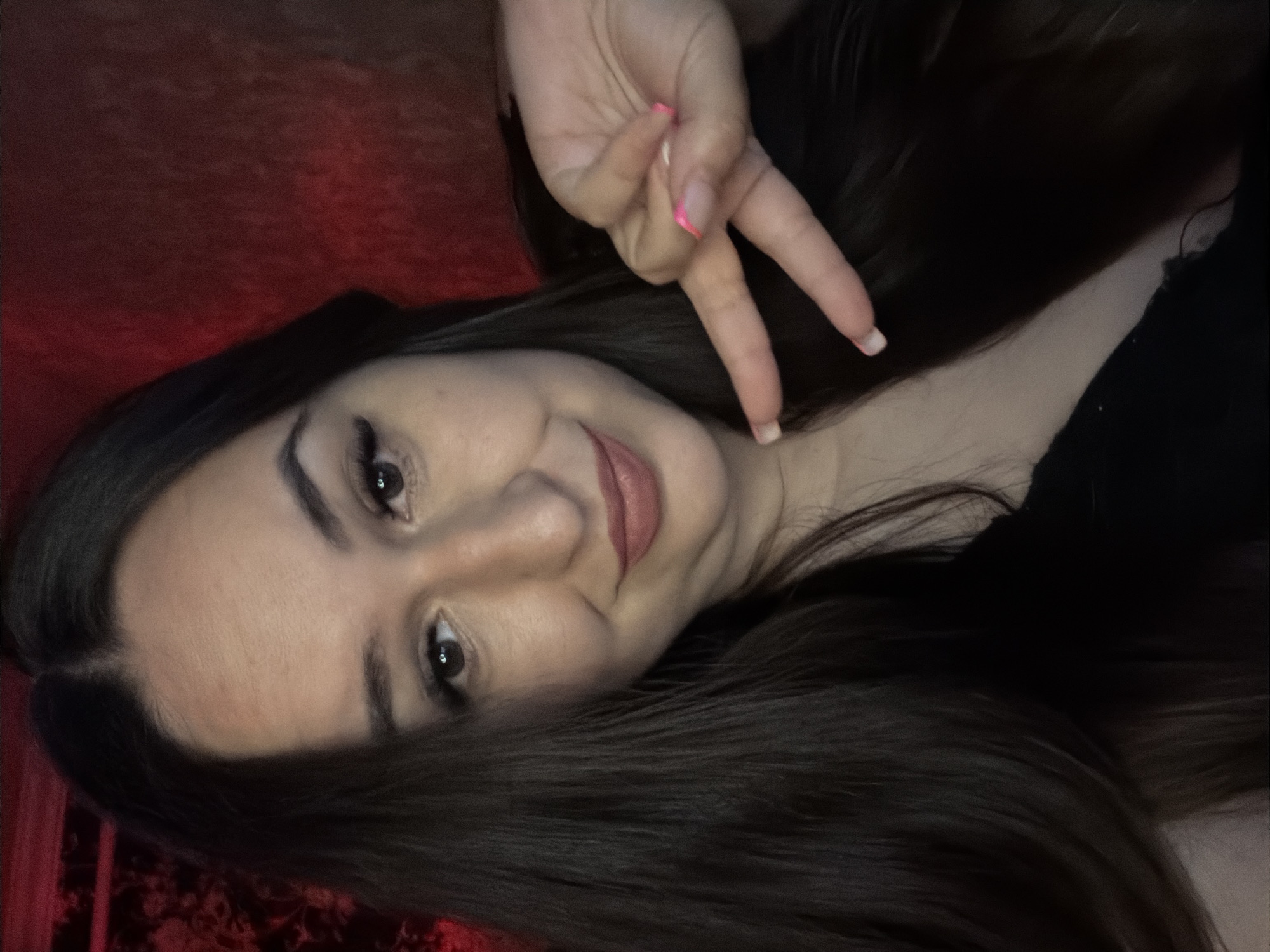 A woman is shown in a close-up, selfie-style shot. She has long, dark hair that is parted in the middle and falls around her face and shoulders. Her eyes are open and looking at the camera, and she is smiling with her lips closed. She is wearing makeup, including mascara and lipstick. Her right hand is raised, and she is holding up two fingers in a peace sign. Her fingernails are painted pink. The background is a dark red, textured surface, possibly a wall or fabric. The lighting appears to be dim, with a reddish hue.