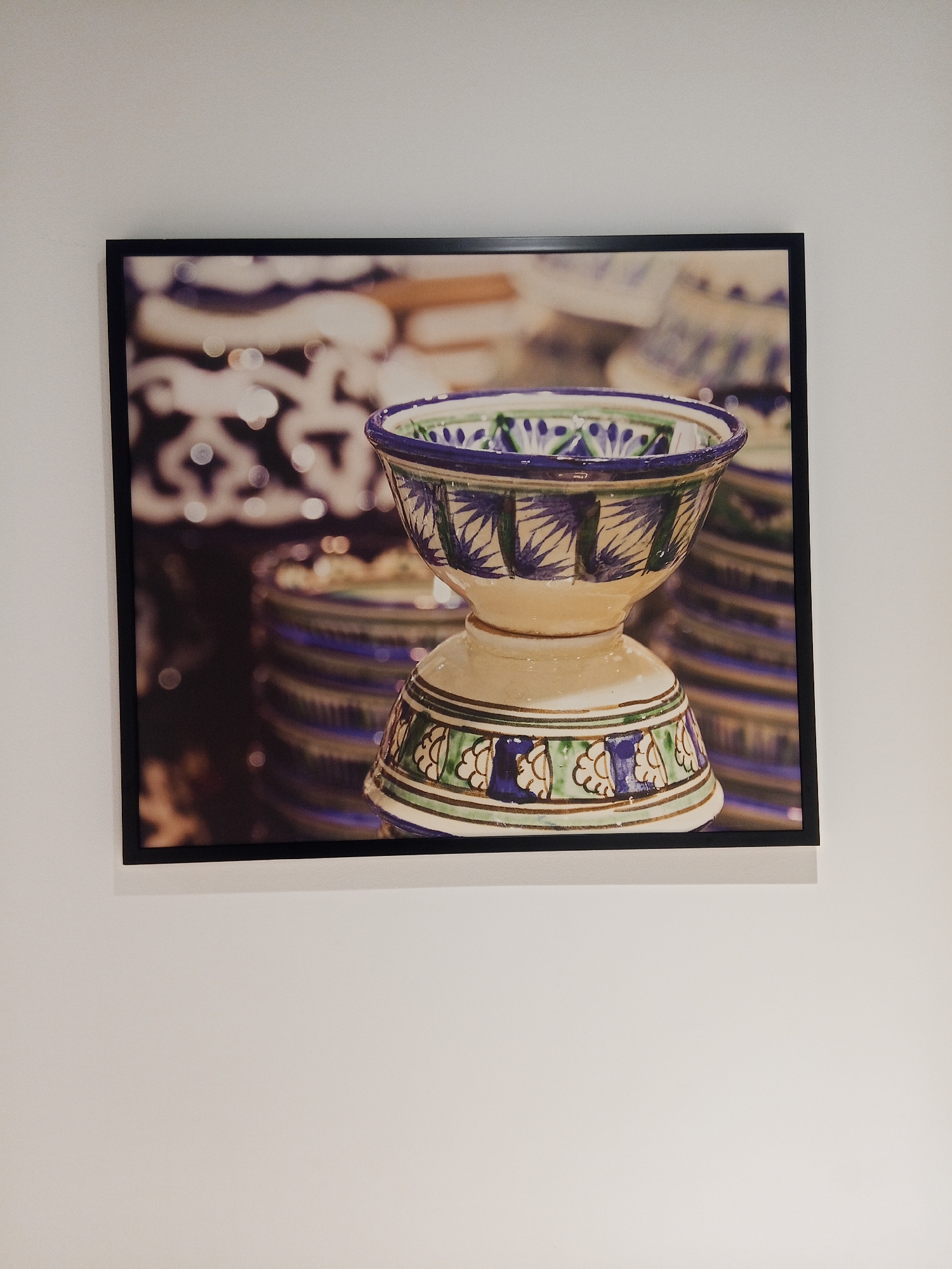 A framed photograph of a ceramic bowl with blue and green floral patterns is displayed on a white wall. The bowl has a wide opening and a pedestal base. In the background, out of focus, are stacks of similar bowls and patterned surfaces. The image is lit from above, casting a slight shadow on the wall below the photograph.