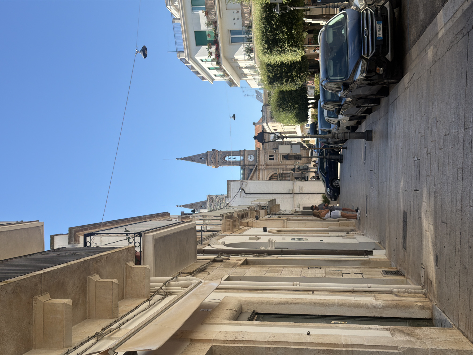The image depicts a street scene in Alberobello, Italy, on a clear, sunny day. On the left, a light-colored building with classical architectural elements, including columns and balconies, lines the street. A woman in a white top and shorts stands on the sidewalk, looking towards a store entrance. Across the street, a row of parked cars, including a black SUV with an Italian license plate FP-766HT, are visible. Further down the street, a white building with balconies adorned with potted plants stands out. In the background, a tall bell tower with a clock face rises above the other structures, indicative of a church or cathedral. The sky is a bright, cloudless blue. The ground is paved with cobblestones, and lampposts are spaced along the sidewalk. Lush green trees are planted along the street, providing shade.