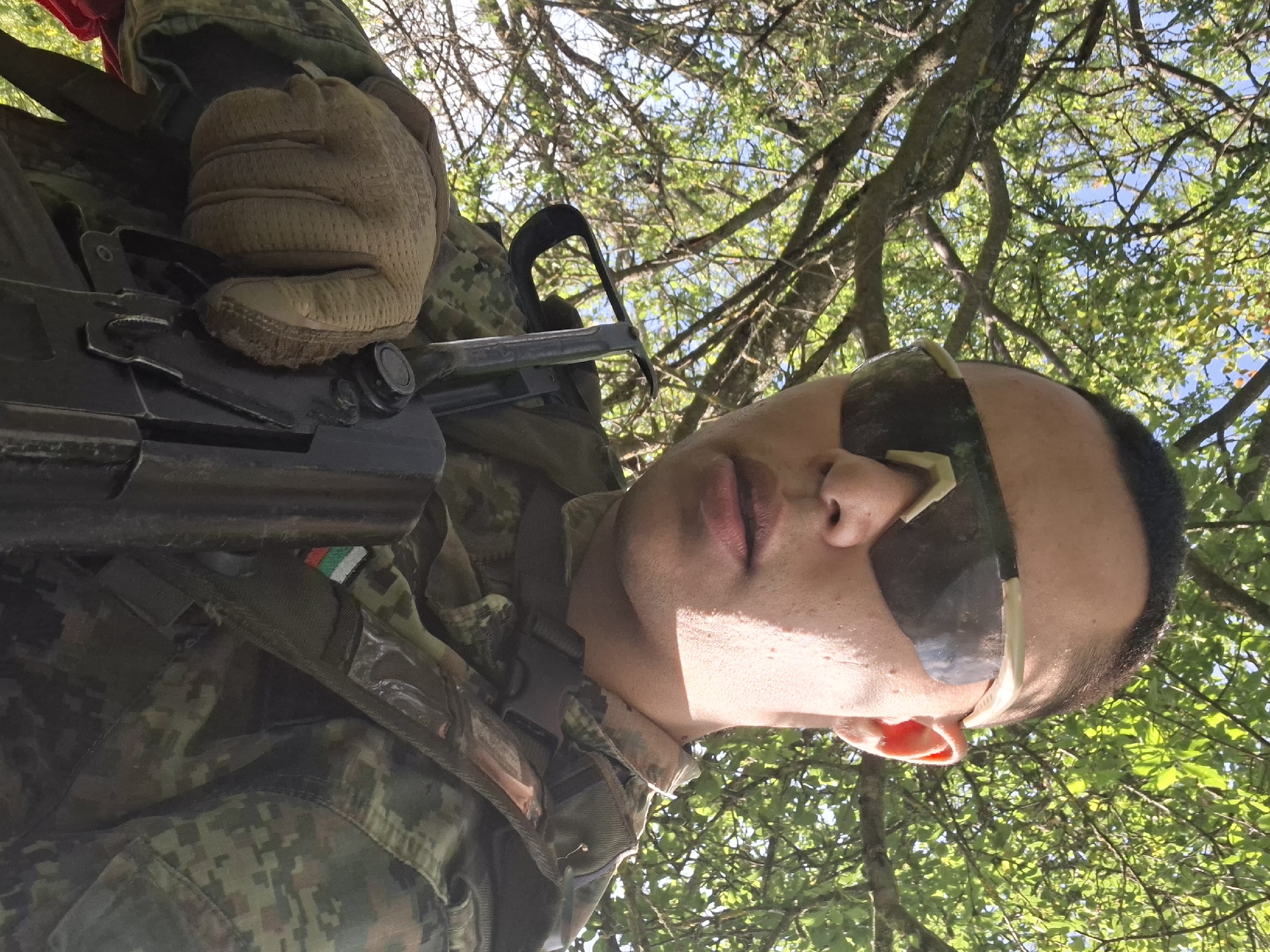 A low-angle, close-up shot shows a person wearing camouflage clothing and tactical sunglasses, holding a rifle. The person has dark hair, light skin, and is looking directly at the camera. The background is filled with green foliage and branches, suggesting an outdoor, natural setting, likely during daytime due to the bright lighting. A Bulgarian flag patch is visible on the person's uniform.