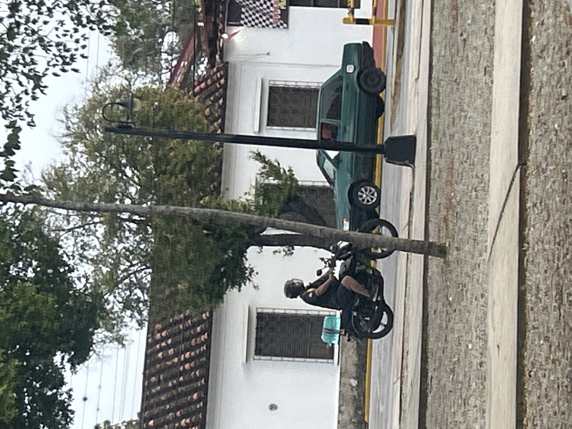 A person wearing a helmet rides a black motorcycle with a blue container attached to the back. The motorcycle is traveling on a paved road with a yellow line. To the left of the motorcycle is a white building with a tiled roof and dark windows. To the right of the motorcycle is a green sedan car. A street light is visible in the middle of the image. The scene appears to be on a street in the city of Santa Ana, Venezuela.