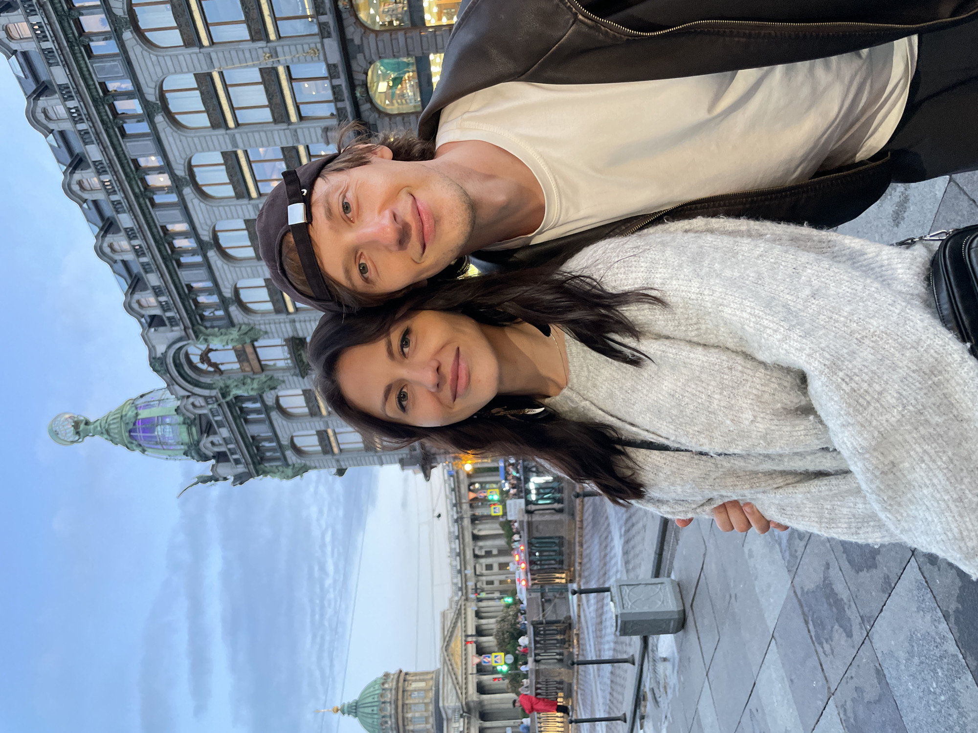 A selfie shows a man and a woman standing in front of an ornate building in St. Petersburg, Russia. The woman, on the left, has long dark hair and is wearing a light gray sweater. The man, on the right, is wearing a black baseball cap, a white t-shirt, and a dark leather jacket. He has light brown hair. Behind them, a grand building with intricate architectural details and large windows is visible. To the left of this building, a large dome with a cross on top can be seen, indicative of Kazan Cathedral. The sky is overcast, suggesting it is dusk or an overcast day. The ground in the foreground is paved with stone. The overall scene depicts a couple posing for a photo in a historically significant urban setting.