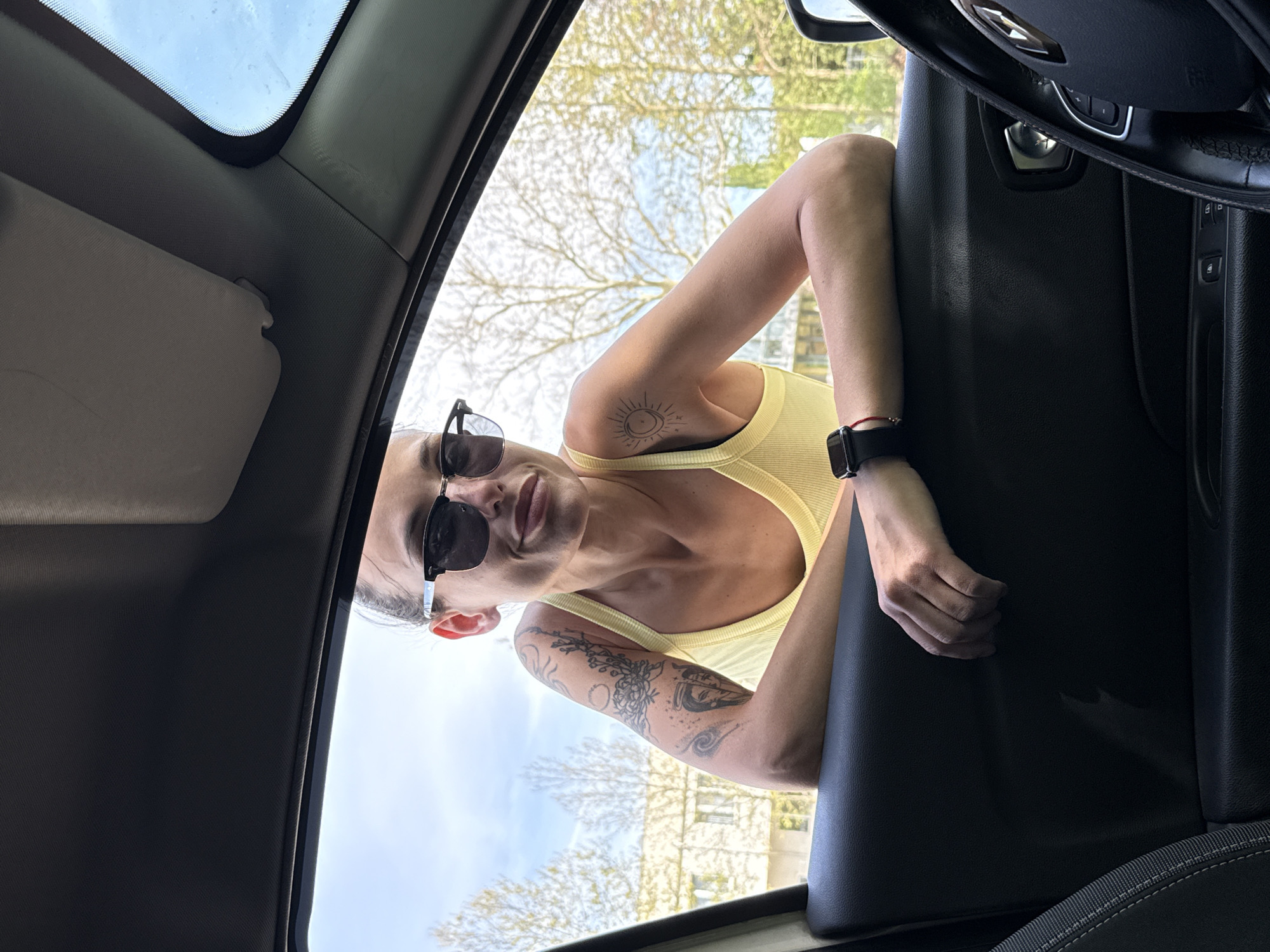 A candid, low-angle photograph taken from inside a car shows a woman leaning into the vehicle from the outside. She is positioned upside down in the frame, wearing a bright yellow ribbed tank top and black sunglasses. Her dark hair is pulled back, and she looks directly at the camera with a slight smile. Tattoos are visible on her arms: a circular, sun-like design on her left shoulder and intricate patterns covering her right forearm. A black smartwatch and a small red bracelet adorn her left wrist.
The car's interior is partially visible, including a gray sun visor, parts of the headliner and sunroof, a dark door panel with controls, and a glimpse of the steering wheel on the right. Through the car's window and above the woman's head, a bright blue sky is visible, indicating daytime. Bare tree branches are prominent in the background, suggesting late winter or early spring, with distant buildings also discernible. The scene takes place outdoors in Pleven, Bulgaria, under sunny weather. No visible text is identifiable.