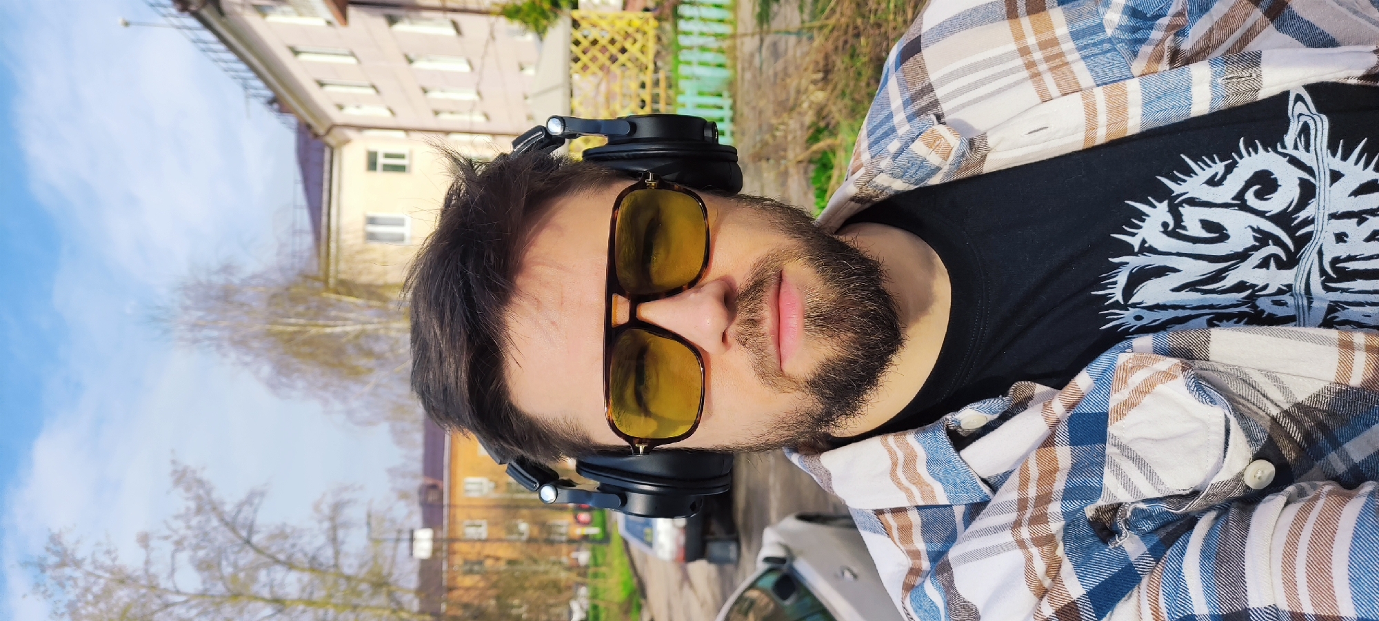 A portrait-oriented, close-up shot features a man outdoors, looking directly at the camera. He has dark hair, a neatly trimmed beard and mustache, and is wearing yellow-tinted aviator-style sunglasses with a dark frame. Black over-ear headphones rest on top of his head and just above his ears.
He is dressed in a black t-shirt adorned with a prominent light-colored graphic design on the chest, which appears to be a stylized creature or abstract pattern with sharp, thorn-like elements. Over the t-shirt, he wears an open plaid flannel shirt featuring a pattern of blue, brown, and white. His lips are slightly parted, giving him a neutral and relaxed expression.
The background suggests an urban or residential street in Fryazino, Russia. Behind him, there's a bright blue sky with some scattered white clouds. Several multi-story buildings are visible, along with bare trees, indicating a late winter or early spring setting. A car is partially visible in the lower right background. The scene is illuminated by bright daylight, suggesting a sunny day. No discernible text is visible on the t-shirt graphic, as it is an artistic design.