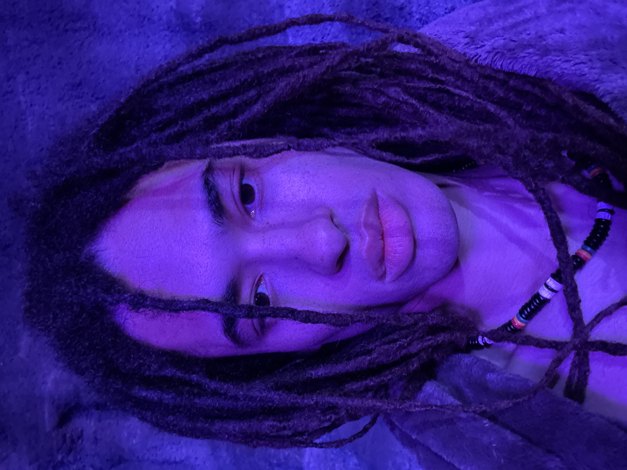 A person with dreadlocks is laying on a surface. The person has light skin and is wearing a necklace with black, white, orange, and green beads. The environment is dimly lit with a blue-purple hue. The location is unknown, and there is no indication that the image was taken in Teguise, Spain.