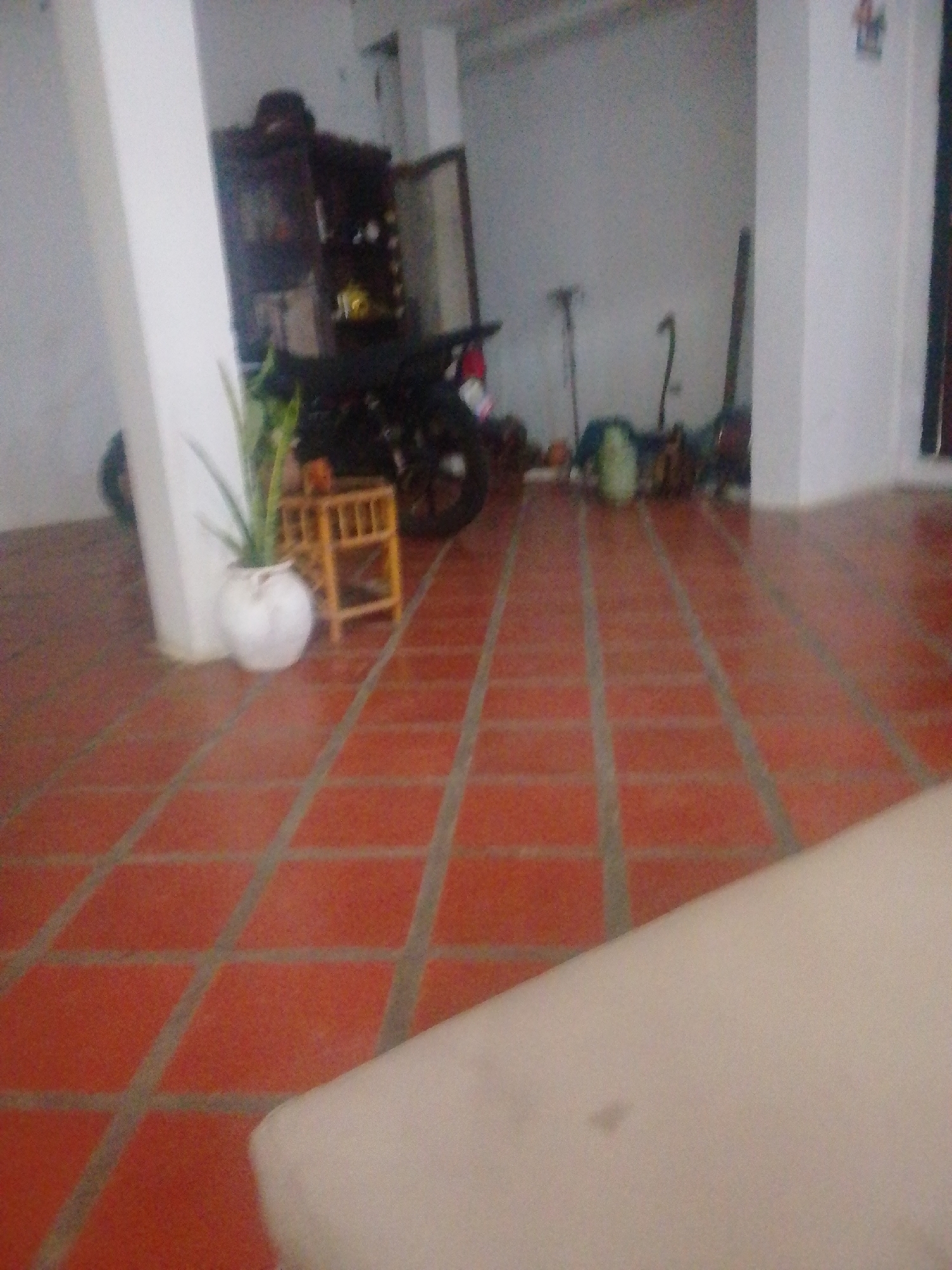 The image depicts an indoor scene with a tiled orange floor. A white pillar is on the left side. In the background, there is a dark wooden cabinet with glass doors. To the right of the cabinet, a motorcycle is parked. A small, wooden stool is next to a white, round vase containing a green plant. Further back, several objects are visible, including what appear to be canes or walking sticks and some green material on the floor. The lighting in the scene is dim. This image is located in Atamo Sur, Venezuela.