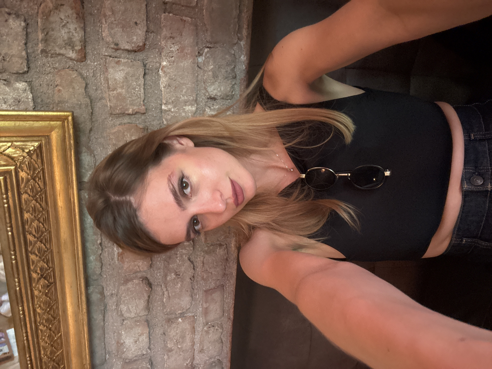 A selfie is taken by a woman with blonde hair and brown eyes, who is wearing a black tank top and denim jeans. She has on makeup and a necklace with a small pendant. Sunglasses are clipped to her shirt. The background shows a brick wall and a gold-framed picture on the wall. The woman is sitting on a dark couch. The scene is likely indoors.
