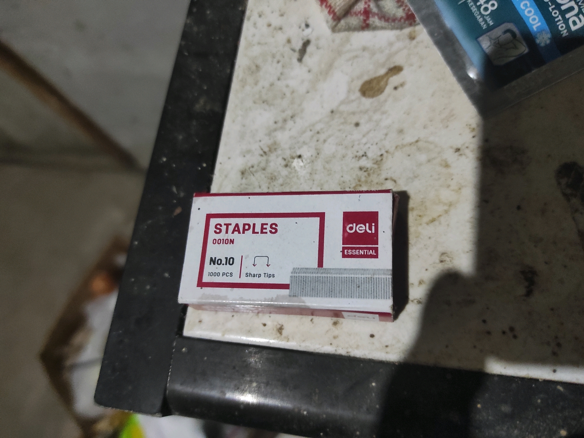 In Buntalan, Indonesia, a box of "STAPLES" branded staples is situated on a speckled, dirt-covered surface. The box is white with red accents, displaying text such as "0010N," "No.10," "1000 PCS," and "Sharp Tips." A "deli ESSENTIAL" logo is also visible. The staples themselves are visible at the bottom right of the box, appearing as a metallic gray row of thin metal fasteners. The lighting casts a shadow on the right side of the image.