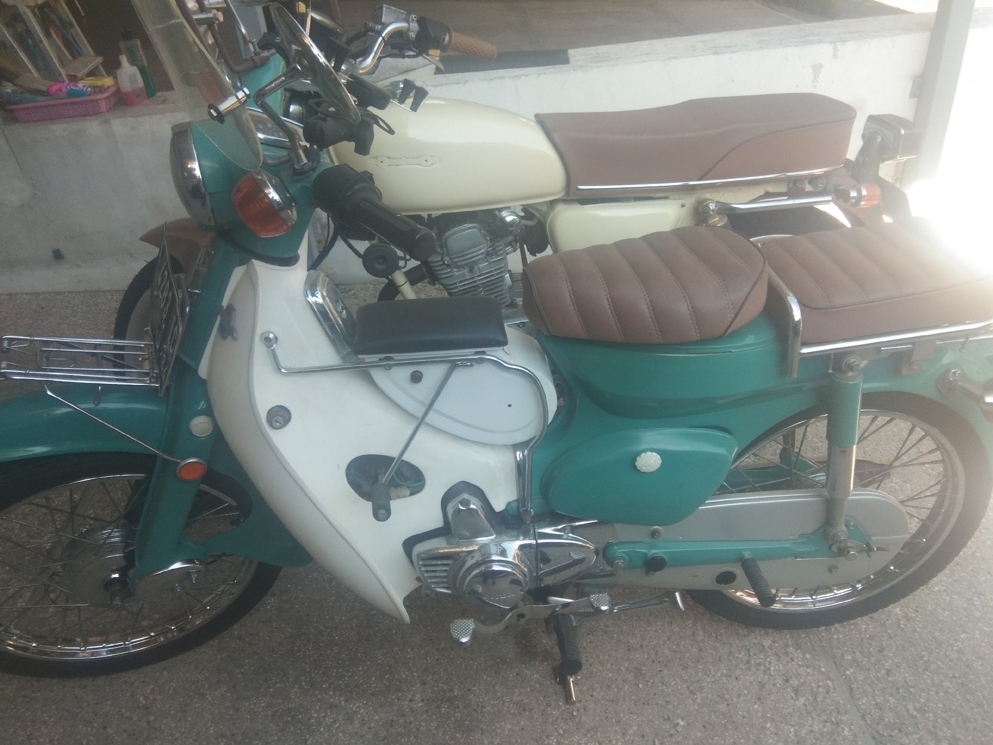 The image shows two vintage motorcycles parked side-by-side on a paved surface. The foreground motorcycle is predominantly white and teal green, with a brown seat and chrome accents. It features a large front fender, a headlight assembly, and a spoked wheel. The engine is visible on the lower left side. The rear portion of the motorcycle shows a teal green fender and another spoked wheel.
The second motorcycle, behind the first, is cream colored with a brown seat and appears to be of a similar vintage style. Its engine is also visible. The motorcycles are parked in what appears to be an outdoor covered area, with a wall in the background. Some shelves with items are visible in the upper left corner of the image, possibly indicating storage or a shop. The lighting suggests it is daytime, with some shadows present. The scene is likely located in Surabaya, Indonesia, given the context provided. There are no people visible in the image.