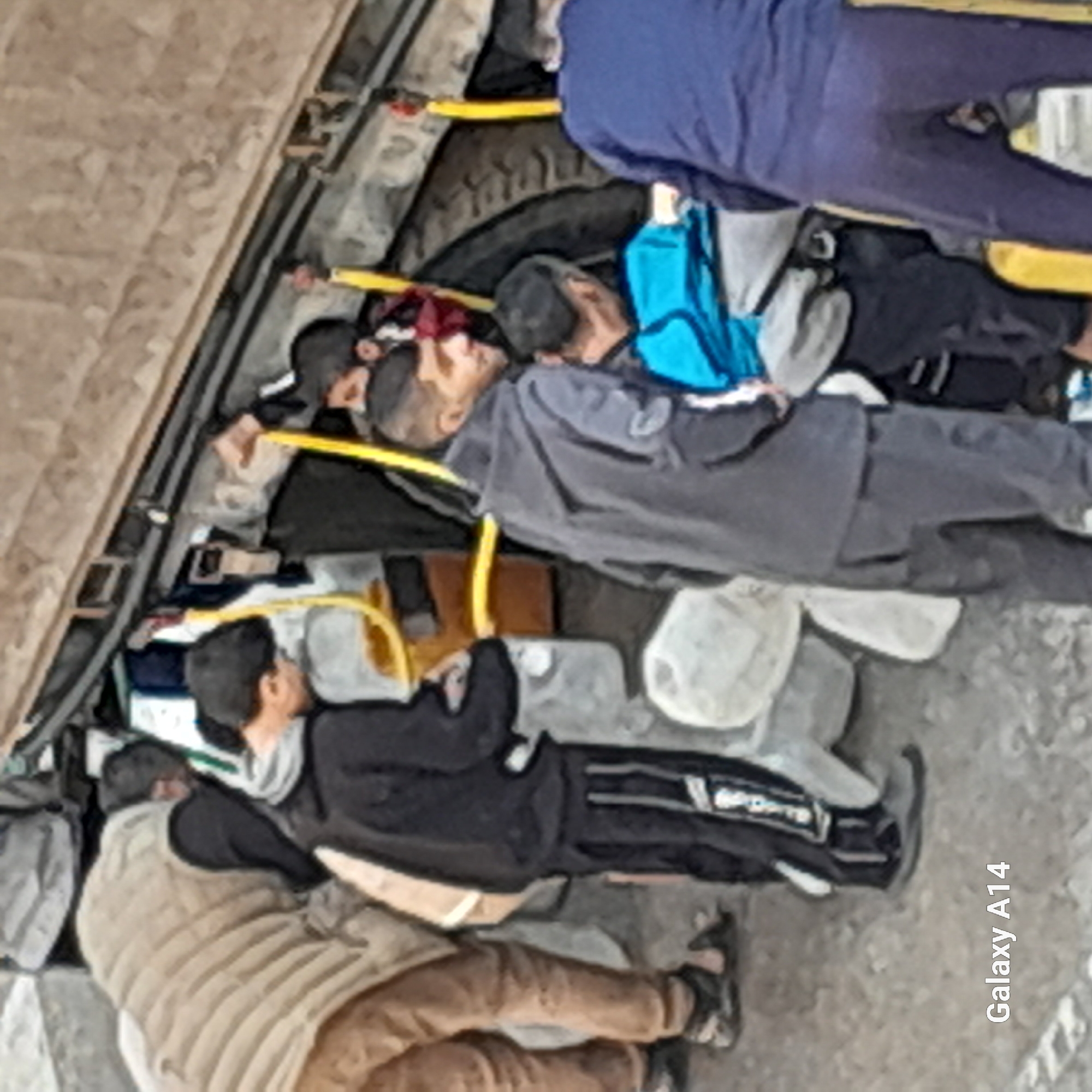 The image shows a group of people lying down, possibly resting or sleeping, in an unknown location in an unknown city. The scene appears to be indoors, possibly a makeshift shelter, with people huddled together on the ground. Some individuals are wearing jackets or vests, and there are personal belongings scattered around, such as bags and clothing. A yellow hose or cord is visible running across the scene.