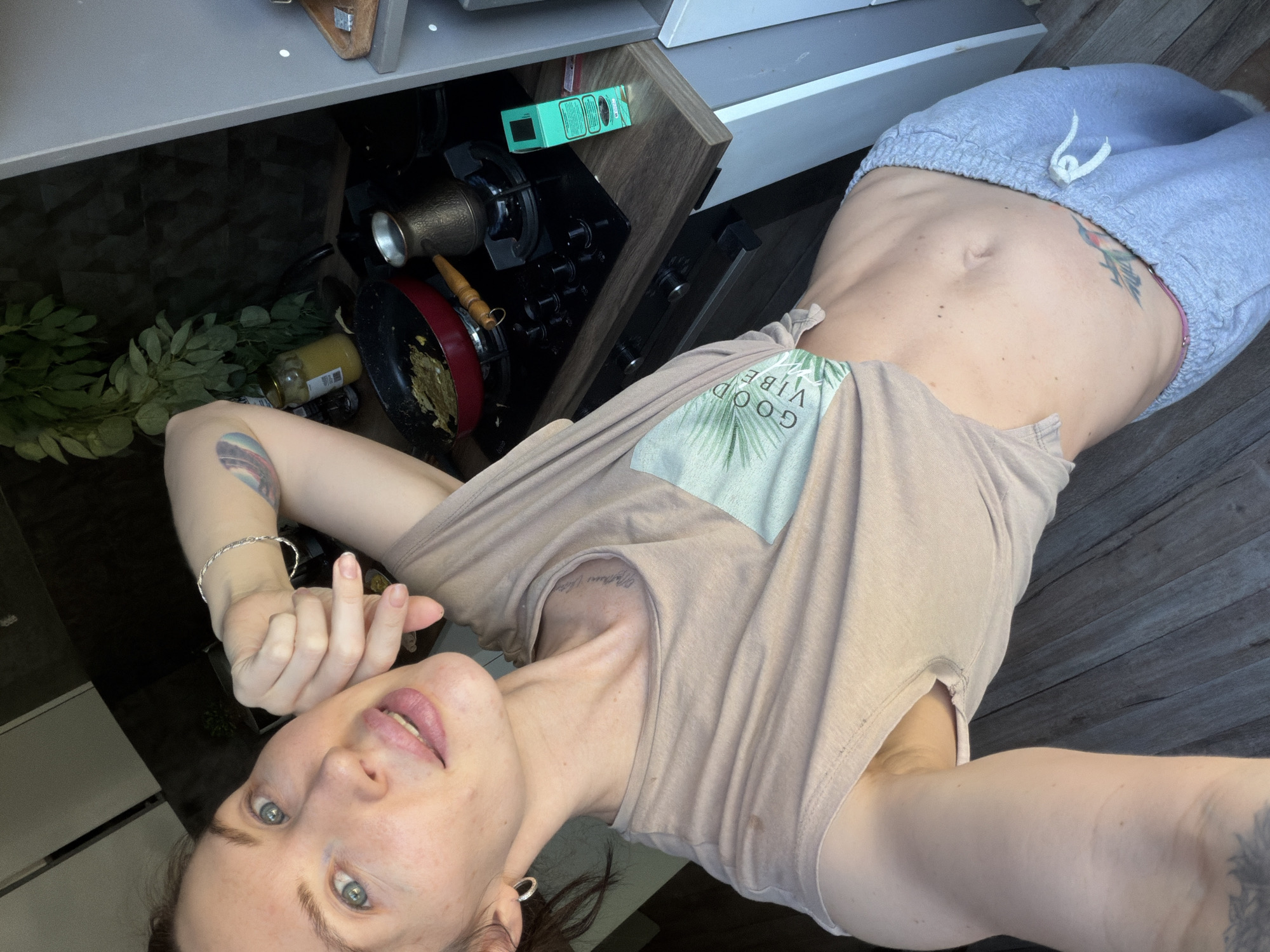 A selfie taken indoors shows a woman with fair skin and blue eyes wearing a beige t-shirt with a palm leaf print and the words "Good Vibes." She has light brown hair pulled back. Her left arm, visible from the elbow down, has a tattoo and a silver bracelet. She's wearing gray sweatpants and has a tattoo on her right abdomen. The background includes a kitchen area with dark cabinets, a stovetop with cookware, and a jar with a yellow substance. The location is within a city in Perm, Russia.