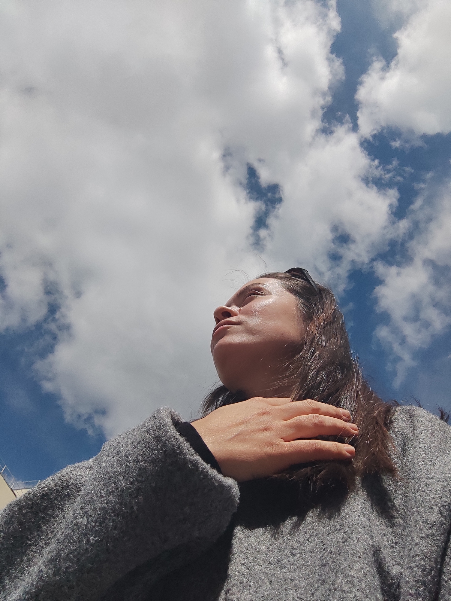 A low-angle shot depicts a person looking up at a sky filled with clouds and blue patches. The person has dark hair and is wearing a gray, textured sweater or coat. Their hand is placed on their collarbone/upper chest area. The sky contains mostly white clouds. A portion of a building is visible. The exact location in Chaikovsky, Russia cannot be determined solely from the details in this image.