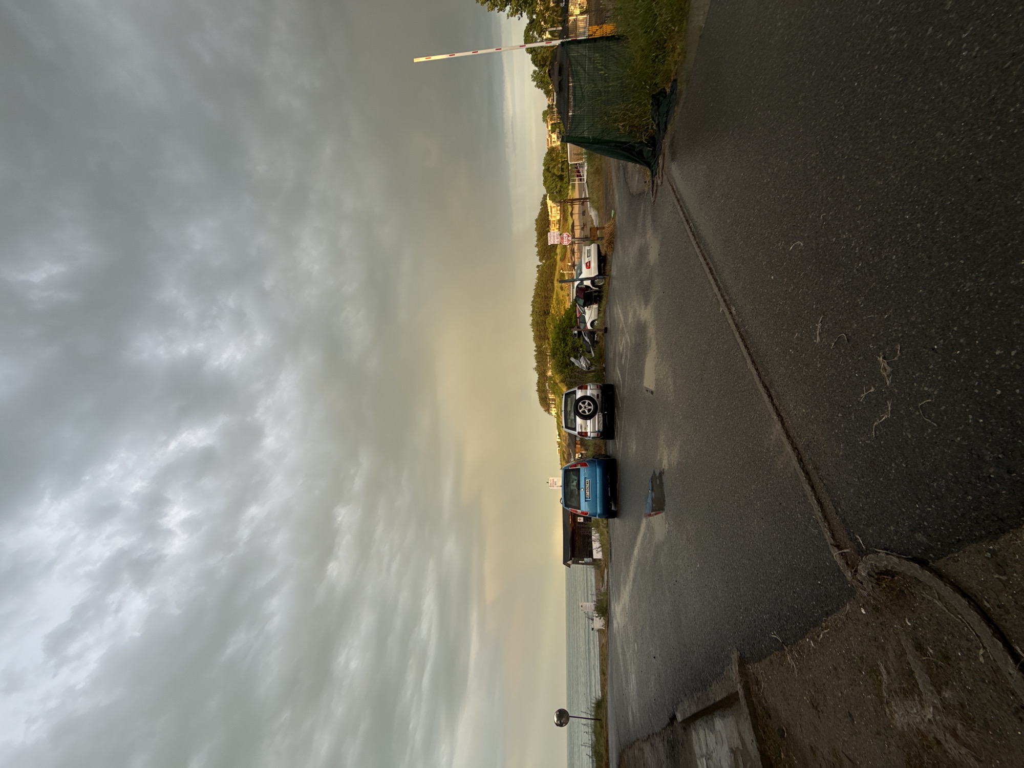 The image depicts a quiet coastal scene under an overcast sky, viewed as if rotated 90 degrees counter-clockwise for proper orientation. The sky, dominating the upper portion, is filled with thick, grey clouds, with a lighter, slightly yellowish hue visible near the horizon on the right, suggesting either early morning or late afternoon light breaking through.
In the mid-ground, a wet, dark asphalt road stretches horizontally, showing puddles and damp patches, indicating recent rainfall. On the right side of the road, several cars are parked; a distinct blue compact car is visible, followed by a silver SUV with a rear-mounted spare tire. Further down the road, more vehicles are partially visible.
To the right of the road and behind the parked cars, the calm, grey-blue surface of a large body of water, likely the Black Sea given the Tsarevo, Bulgaria location, extends to meet the cloudy horizon. On the left side of the road, there's a strip of green grass and foliage, leading to dense trees and some distant buildings or houses nestled within the greenery.
A prominent white and red striped boom barrier is visible on the left side, extending over what appears to be an access path or entrance. Nearby, a red octagonal "STOP" sign is clearly visible. The blue car's license plate shows "BG" (for Bulgaria) and some partial numbers. No people or overt activities are present, lending a serene, post-rain atmosphere to the scene.