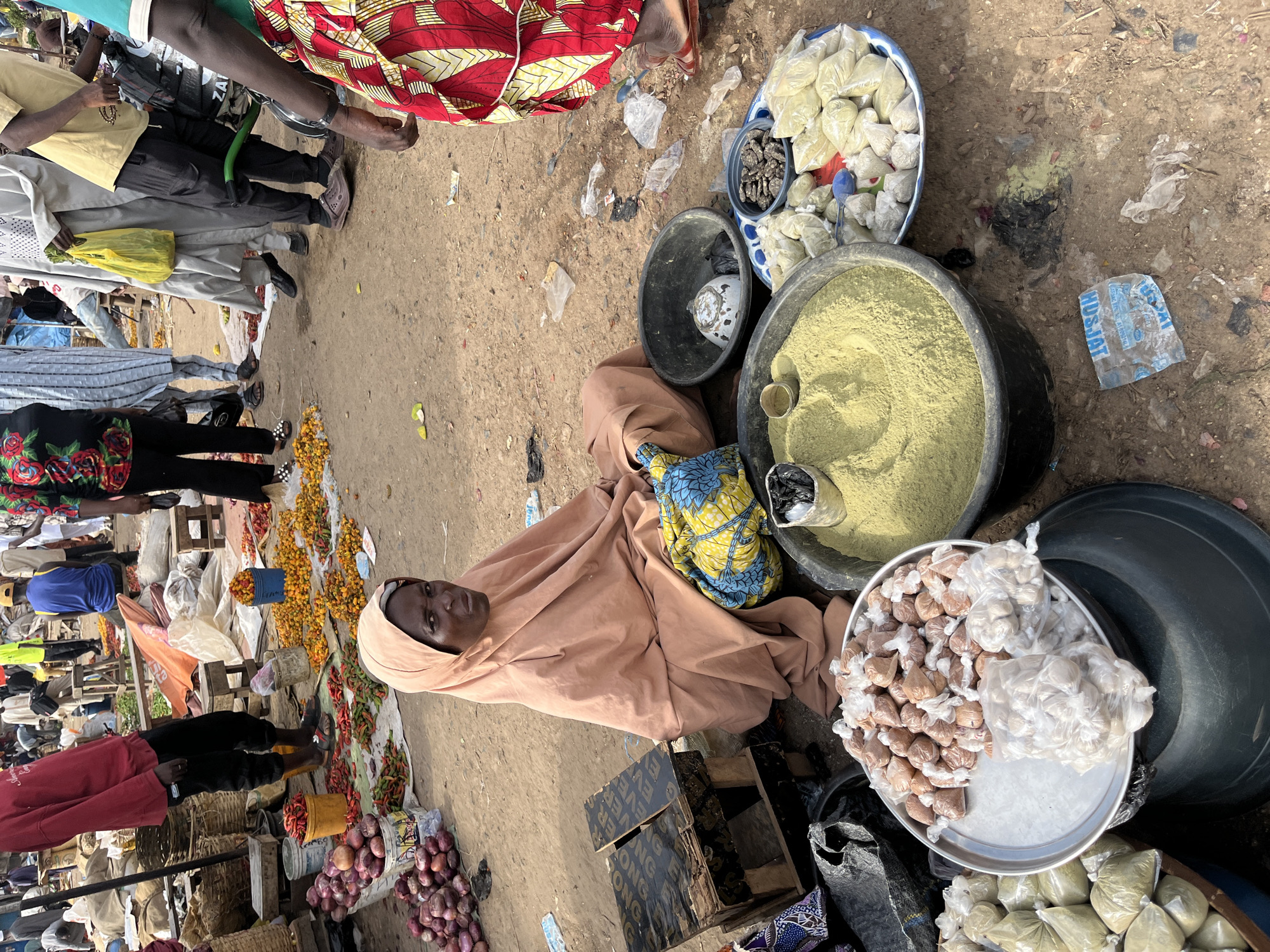 The image shows a busy outdoor market scene in Rafin Par, Nigeria. A woman wearing a peach-colored hijab and traditional clothing sits on the ground next to a display of various goods. In front of her, there is a large black basin filled with a yellow powder, likely spices or ground grains. To her left, a silver platter holds many small, individually wrapped items, some in clear plastic and others in light brown paper, appearing to be packaged food items or dried goods. Next to this platter are several clear plastic bags filled with a light green substance, possibly spices or herbs. To the right of the yellow powder basin, a small bowl contains dark, irregularly shaped objects, possibly dried seeds or spices. Behind the woman, a colorful patterned fabric is draped, and a black plastic bag sits nearby. The ground of the market is dirt and sand, with scattered debris including plastic wrappers and leaves. In the background, other market-goers are visible, some standing and others walking, with stalls and more produce, including red onions and red and yellow chili peppers, laid out on the ground. The overall impression is one of a vibrant, active marketplace.