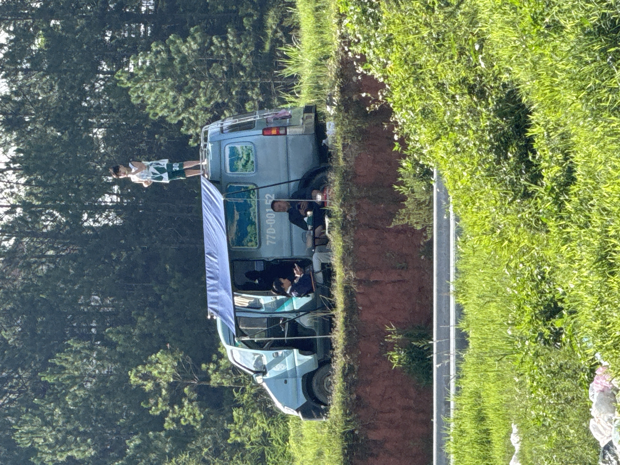 This image captures a roadside scene in Da Lat, Vietnam, under bright daylight. A light blue minivan is positioned on a grassy, reddish-brown dirt embankment, elevated above a partially visible paved road and a grey concrete barrier in the foreground. A dense forest of tall evergreen trees forms the background.
On the roof of the van, a young child, barefoot and wearing a white and green sleeveless top with green shorts (numbered "30"), stands upright, facing towards the left and appearing to hold small items in his hands. A blue fabric awning extends from the van's right side, providing shade. Beneath this awning, two adults are seated. One adult, with dark hair and wearing a dark top, is seated in a portable folding chair outside the van's open sliding door, focused on a white object (possibly a book or device) held in her hands. The second adult, dressed in dark attire, is seated directly on the ground beside the van's rear right wheel, looking down at a black smartphone. A red cup is visible next to this individual.
The van's side displays the alphanumeric sequence "770-001 52" and features decorative cloud-like motifs on its rear windows. A ladder is attached to the rear of the vehicle. The immediate foreground comprises vibrant green grass and some scattered light-colored refuse. The scene suggests a leisure stop or temporary encampment.