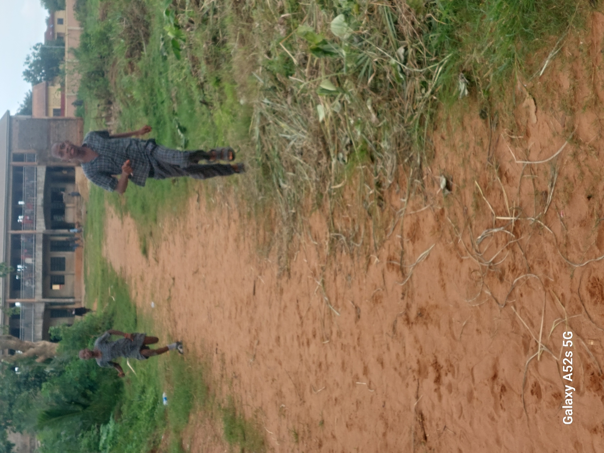 Two young boys are running on a dirt path in Umudioka, Nigeria. The boy in the foreground, wearing a checkered shirt and pants, is running towards the camera. He is wearing sandals. The second boy, in the background and to the left, is also running away from the camera. He is wearing a short-sleeved shirt and shorts. In the background, there is a two-story building with balconies and windows. The area around the path is covered in grass and dirt, with some vegetation visible. The sky is overcast.