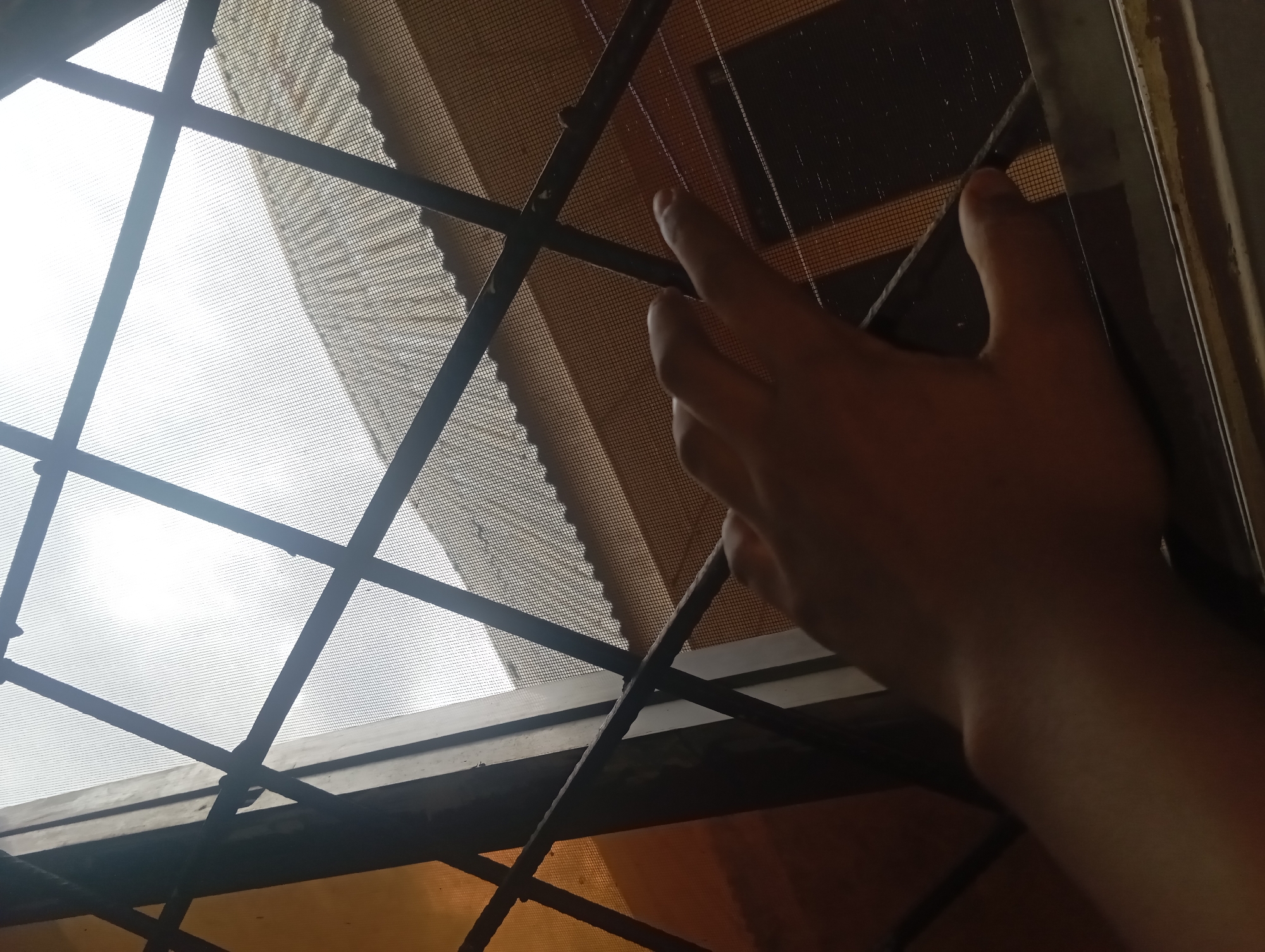 A person's hand is visible reaching towards a window covered by a screen and security bars. The hand appears to be a right hand, with fingers extended and touching the screen. The background shows the exterior of a building with a tiled roof, partially obscured by the window screen and bars. The sky is overcast. The scene appears to be an interior view looking outwards through a window.