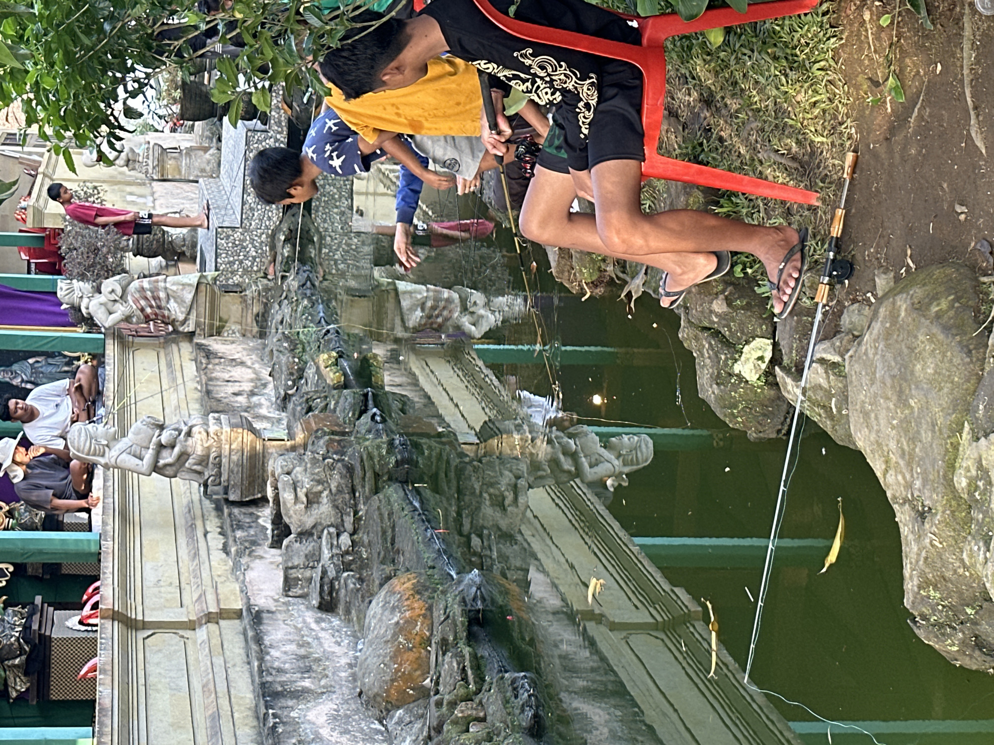 In Bangli, Indonesia, two boys are fishing in a pond. The boy on the left is wearing a blue shirt with white bird patterns and grey shorts. He is leaning over the pond, looking into the water with a fishing rod in his hand. The boy on the right is wearing a black long-sleeved shirt with gold designs and black shorts. He is sitting on a red plastic chair, also holding a fishing rod and looking towards the pond.
The pond has green, murky water, with rocks and some fallen leaves on its surface. Several fishing rods are visible, cast into the water. The background shows a building with a stone facade and decorative statues, suggesting a temple or a traditional complex. Trees and lush greenery are also present, indicating an outdoor setting. The lighting suggests it is daytime, possibly late afternoon, with sunlight casting reflections on the water.