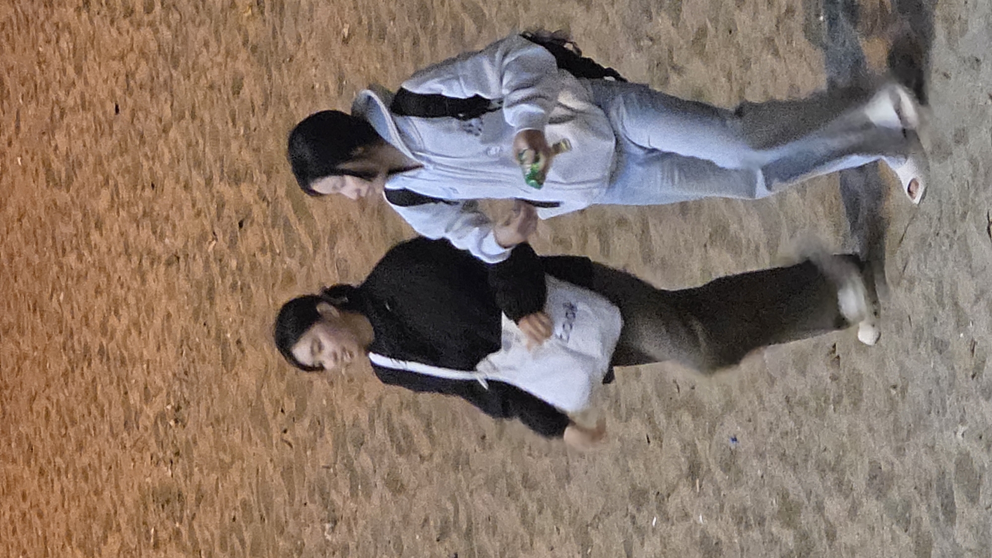 Two individuals, both likely young women, are walking on a sandy surface, possibly a beach. One is wearing a light-colored hooded jacket, light-colored pants, and carrying a dark-colored backpack. She appears to be holding a green object in her right hand. The other person is wearing a dark-colored jacket with a white stripe, dark pants, and carrying a white bag with text that is partially visible. The ground is sandy, suggesting an outdoor setting. There are no obvious landmarks to confirm the location in Nha Trang, Vietnam, but the sand suggests a coastal area. The image appears to be taken from above, looking down at the individuals.