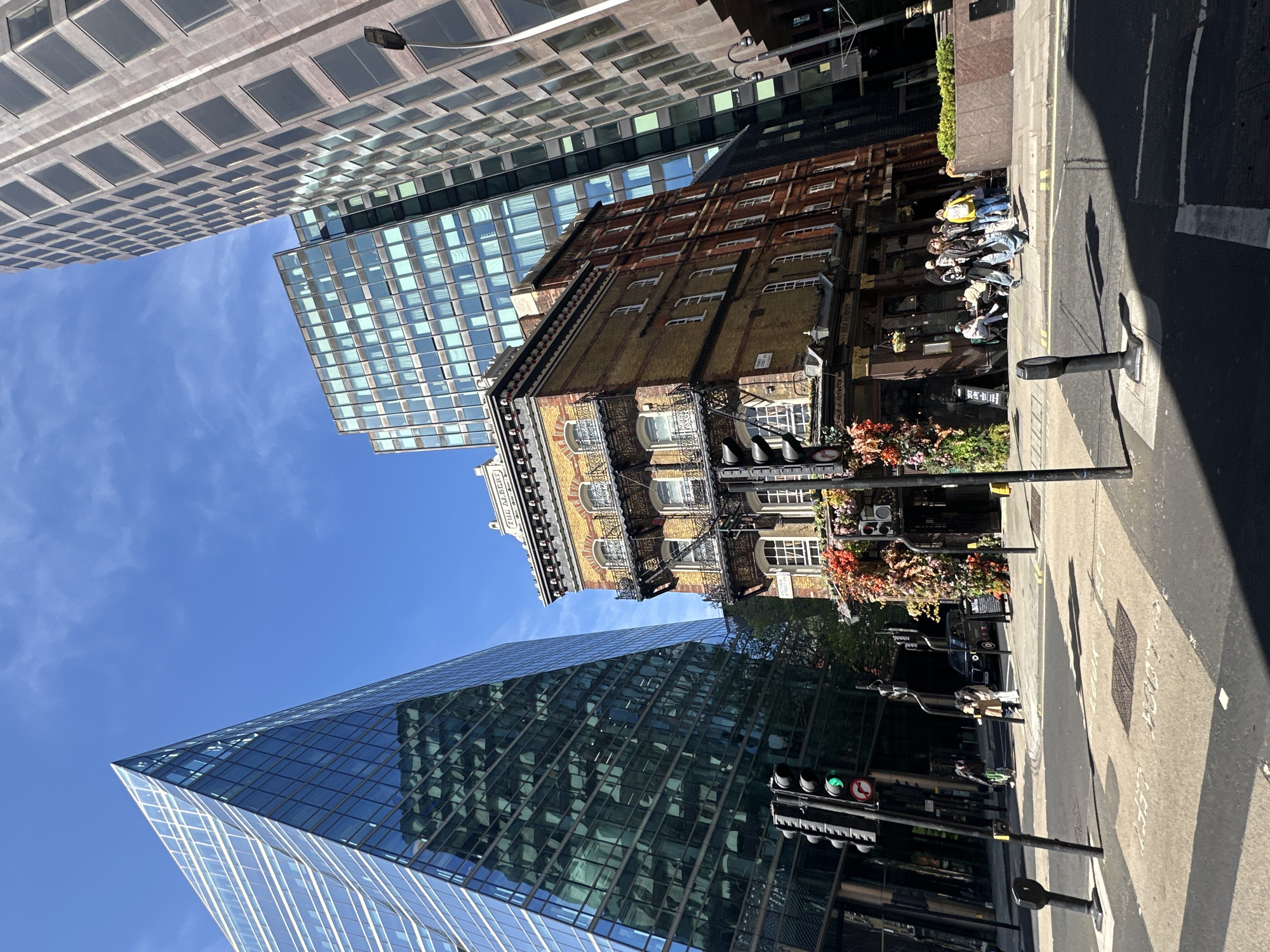The image captures a street scene in London, United Kingdom, showcasing a blend of modern and traditional architecture. A modern glass skyscraper dominates the lower foreground on the left. Above and slightly to the right stands a yellow brick building with visible architectural details and a sign that reads, "The Albert." Above that is a newer skyscraper. On the right side of the image, people are seated at outdoor tables in front of a restaurant or pub, possibly enjoying a meal or drinks. Pedestrian traffic can be seen on the sidewalk. Traffic lights are visible, showing a green light. The street pavement is visible, with visible manhole covers. The sky above is blue with few clouds. The scene appears to be shot from a low angle, tilting upwards to emphasize the height of the buildings.