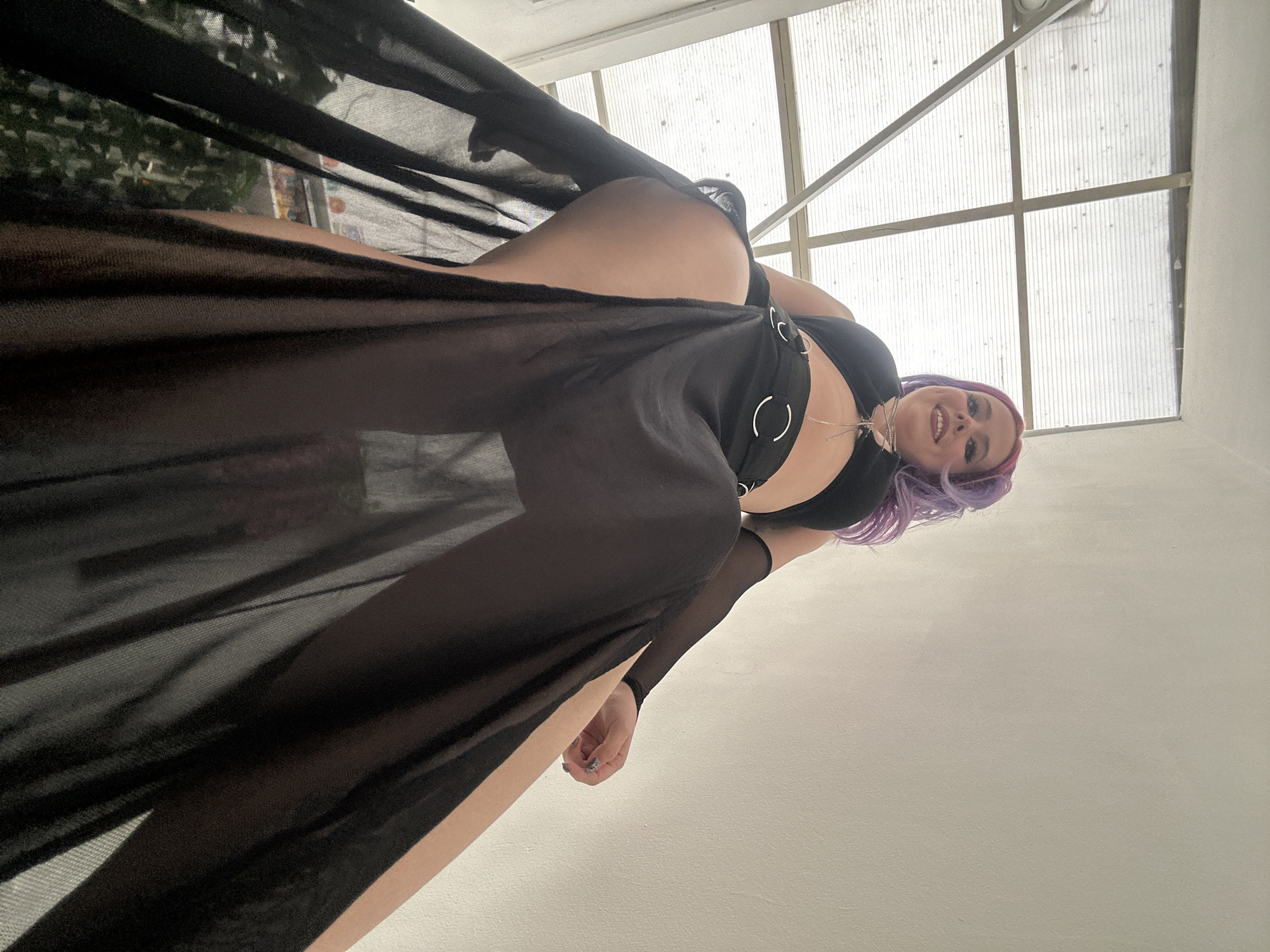A low-angle shot shows a woman with purple hair wearing a black outfit with circular accents and a sheer black overlay. She is looking down at the camera and smiling. The background includes a ceiling with a grid of translucent panels. Based on the location information, this could be a building in the city of Zagreb, Croatia.