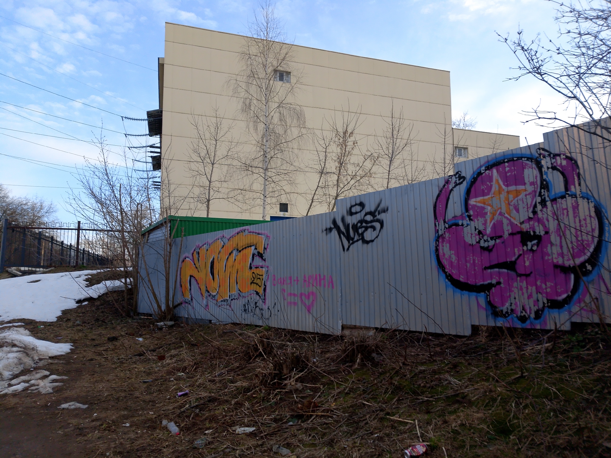 The image shows a section of a corrugated metal fence covered in graffiti, located in what appears to be a neglected area in Izhevsk, Russia. The fence is adjacent to a brown, grassy area with patches of melting snow. Behind the fence is a large, plain beige building with a few visible windows. Several bare trees and bushes are interspersed between the fence and the building. A secondary fence is visible in the background on the left side of the frame. Power lines are strung across the upper-left portion of the sky, which is partly cloudy. The graffiti on the fence consists of various colorful designs and words. Debris litters the ground.