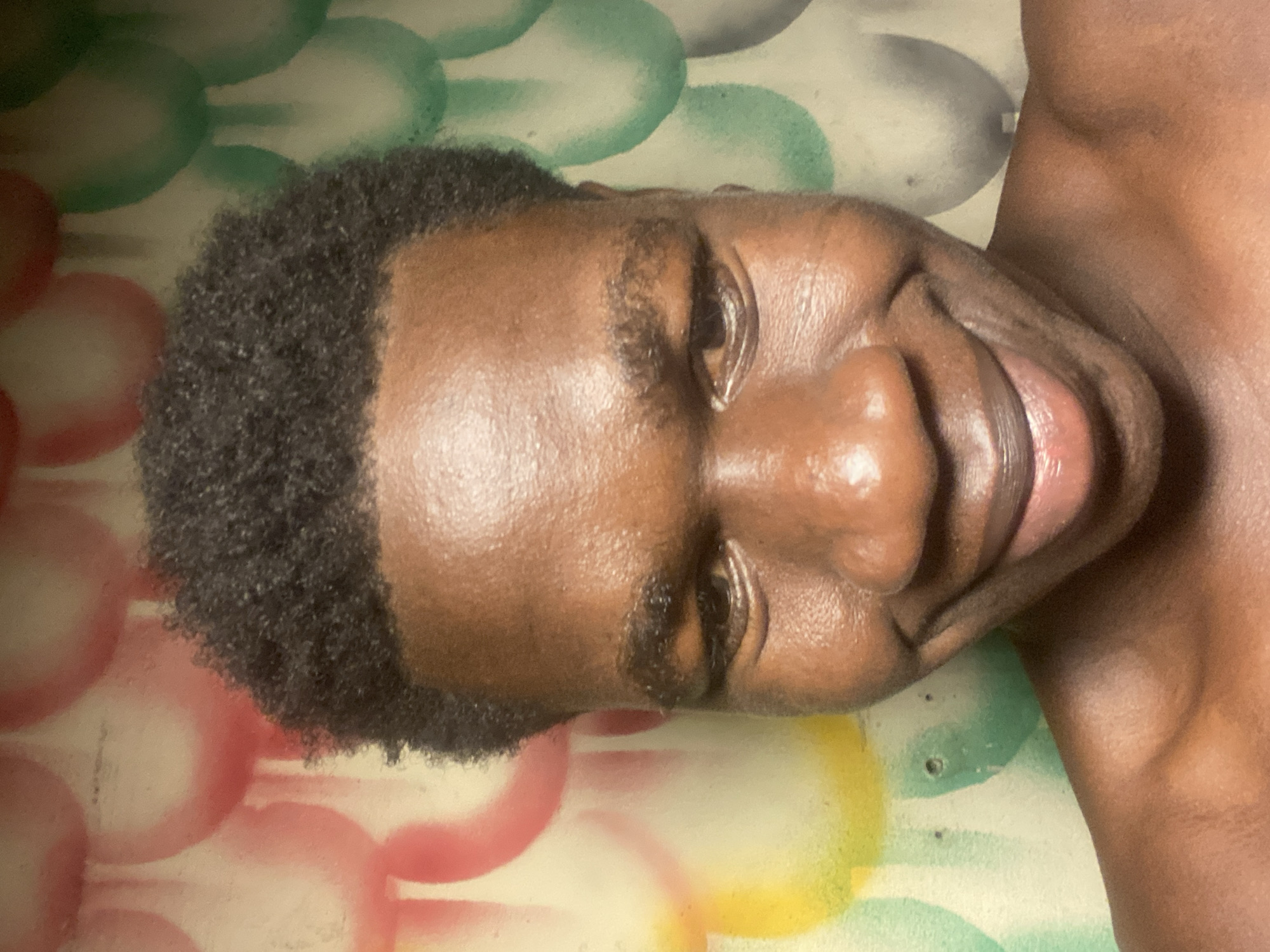 An eye-level, close-up portrait of a dark-skinned man lying on a surface with a colorful spray-painted design. He is smiling slightly, with his head tilted. His hair is short, dark, and curly. The surface he is lying on is painted with intersecting circles of red, green, gray, and yellow. The man's chest is visible in the upper right corner. Location information is not determinable from the visual information provided.