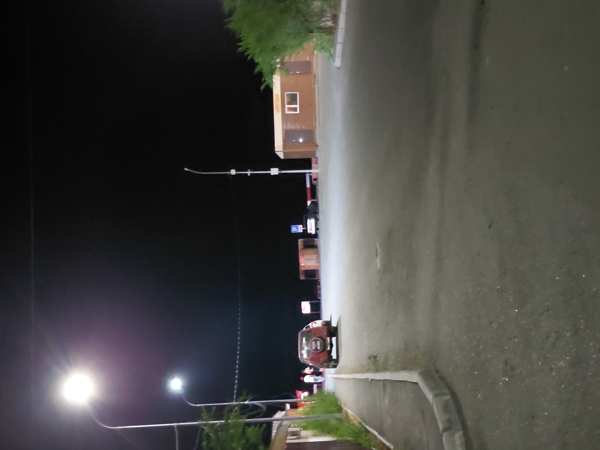 The image depicts a street scene at night in Ақши, Kazakhstan. A red SUV is parked on the right side of a paved road, facing away from the camera. To the left of the SUV, a sidewalk runs alongside a low-rise building. Further down the sidewalk, a group of three people are walking away from the camera. To the right of the road, there is a small wooden building with a white-framed window and two doors. A street light illuminates the scene from the upper left. In the background, dark foliage and a dark sky are visible. The overall impression is of a quiet, possibly rural or suburban area at night.