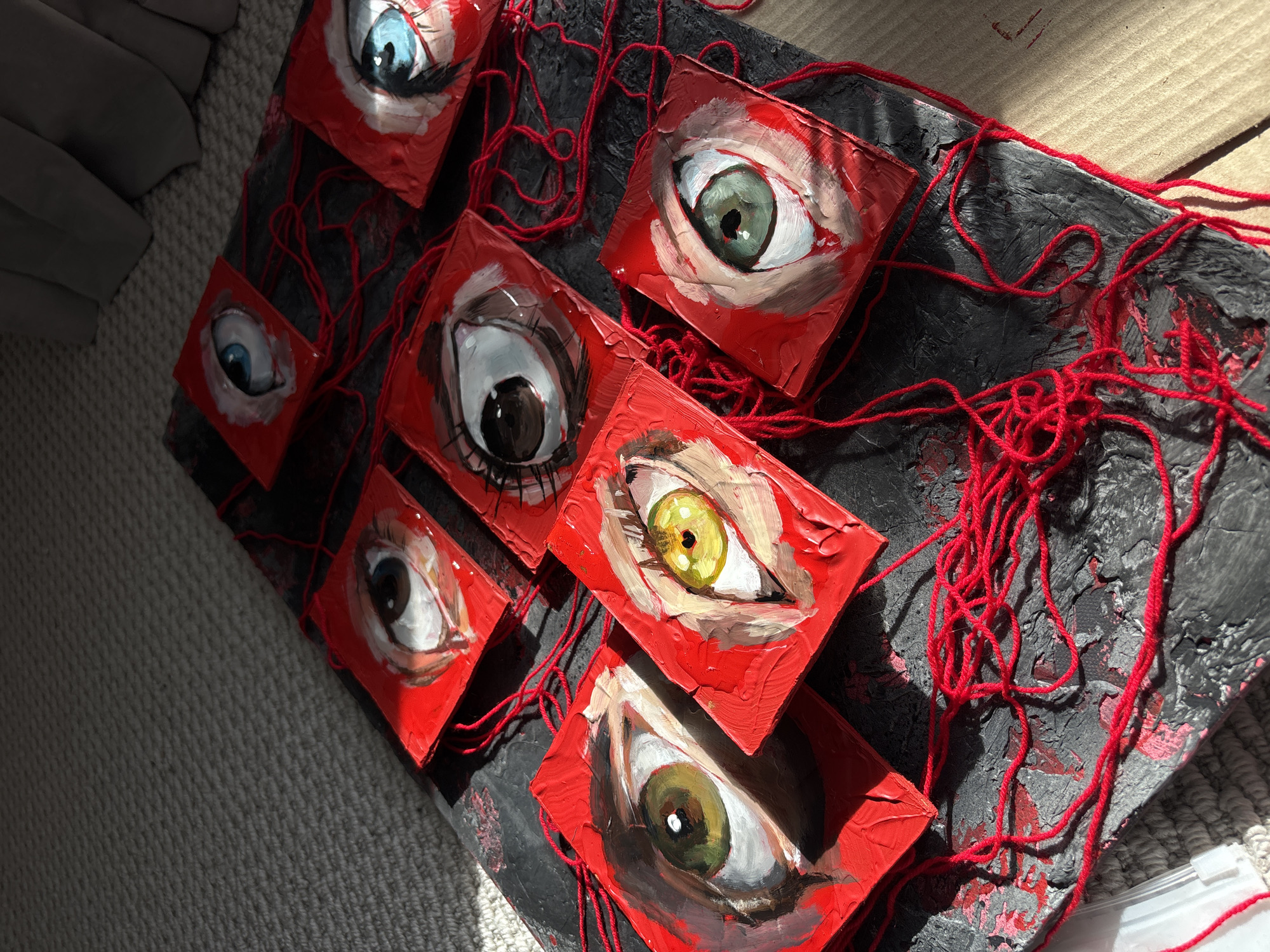A slightly high-angle close-up shot shows a collection of small square paintings of eyes, arranged on a dark, possibly painted, textured surface and connected by red thread. The paintings are primarily in shades of red and white, with varying eye colors. The background surface has a rough, distressed appearance, with visible cracks and color variations. Red thread is draped across the surface, connecting the paintings and adding a sense of interconnectedness or tension. The overall lighting suggests a mix of natural and artificial light sources. The bottom right of the shot shows a glimpse of a white plastic box and what may be flooring. There are no discernible landmarks to tie the image to Krasnoyarsk, Russia. No people are visible.