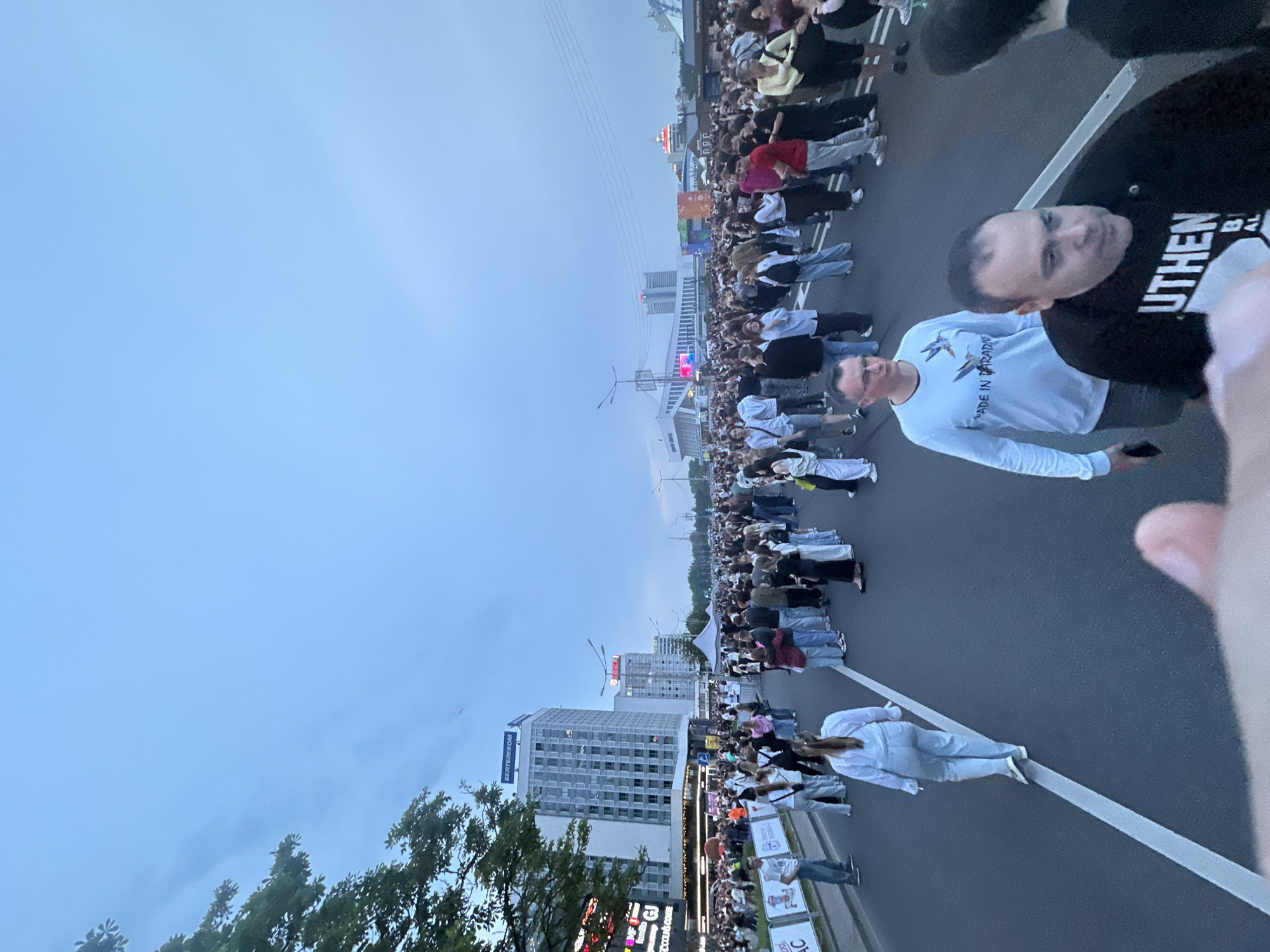 This image depicts a large public gathering on a multi-lane city street in Minsk, Belarus, likely during dusk or an overcast day. The street is densely populated by hundreds of individuals, predominantly young to middle-aged adults, dressed in casual attire, with many wearing white, black, or light-colored clothing. They are standing and walking in a generally uniform direction, occupying the entire width of the asphalt road, which features prominent white lane markings.
In the immediate foreground, a male individual wearing a light blue long-sleeved shirt with a "MADE IN PARADISE" graphic featuring two hummingbirds walks towards the viewer's right, holding a dark object in his left hand. Partially obscuring him and in closer foreground is another male individual in a black hoodie with partially visible white text reading "UTHEN B AL". A woman in a white long-sleeved shirt and light blue pants walks away from the viewer along a white lane marking in the mid-ground. A blurry hand is visible in the bottom-center foreground.
To the left, multi-story urban buildings line the street. A prominent modern high-rise features a large "БЕЛТЕЛЕКОМ" (BELTELECOM) sign on its roof, and various commercial storefronts are visible below. Metal barriers with banners (partially reading "AC") separate the crowd from the sidewalk area on the left. In the distance on the right, several more buildings are visible, including a large, modern white building with a distinctive architectural roofline, possibly a sports or event arena, and other structures with red illuminated signs. White event tents are set up on the right side of the street. Overhead utility lines stretch across the sky in the middle-right. Green foliage from trees is visible on the far left and in the distant right. The sky is overcast with a light blue-grey hue. A small, faint red object, possibly a kite or balloon, is visible in the upper right sky.