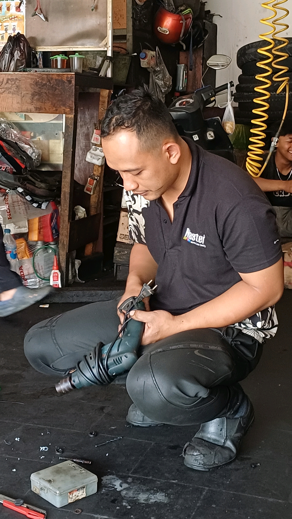 A man wearing a black polo shirt with a logo and black pants is squatting on a dark tiled floor in what appears to be a workshop or garage. He is holding a green power drill with a black cord. Several small objects are scattered on the floor around him, including what looks like screws, nuts, and a small toolbox. In the background, there are shelves with various items, including bottles of liquid and plastic bags. A red motorcycle helmet is visible hanging on the wall. To the right, another person is partially visible, seated and smiling, with a yellow coiled hose behind them. The overall setting suggests a place where repairs or maintenance are performed.
