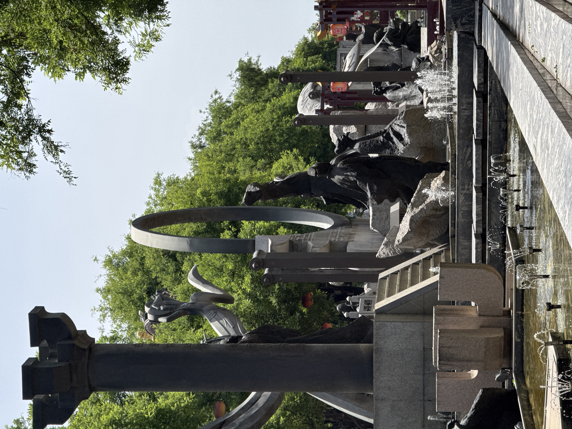 The image depicts a monument in Yanta District, China. The structure is a series of layered stone formations and sculptures. There are gray metal arches and columns. Water is cascading down the stone formations into a pool. Lush green trees are visible in the background. The sky is pale blue.