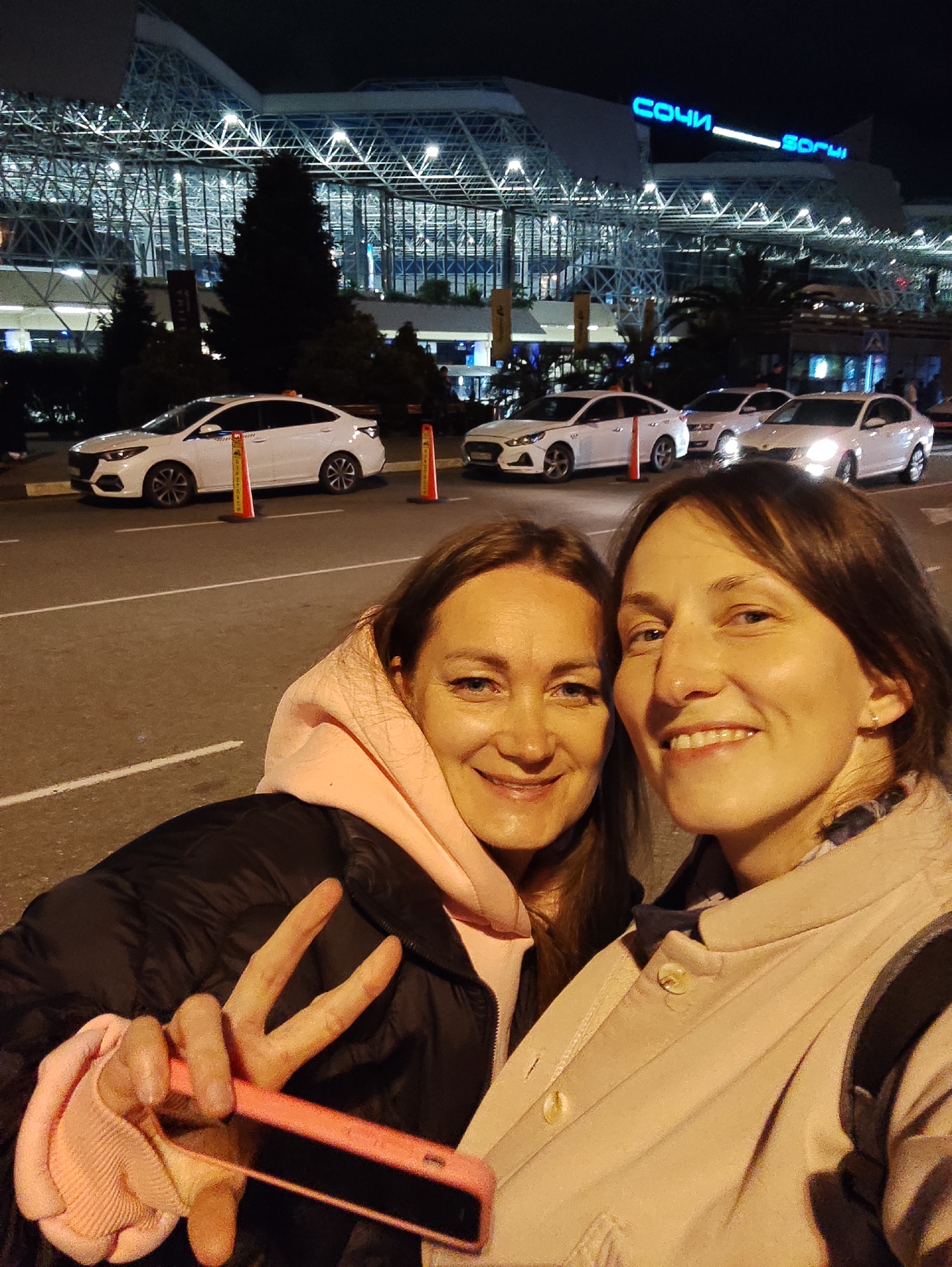 The image presents two adult females in the foreground, positioned for a selfie-style photograph taken at night. The setting is identified as Sochi International Airport in Sochi, Russia, evidenced by the illuminated "СОЧИ" (Sochi in Cyrillic) and "SOCHI" signs prominently displayed on the main terminal building in the background.
The woman on the left, with brown hair and blue eyes, is wearing a light pink hooded sweatshirt beneath a dark, possibly black, puffer jacket. She holds a pink-cased smartphone in her left hand and is making a V-sign (peace sign) with her index and middle fingers. The woman on the right, also with brown hair and blue eyes, wears a light beige or pale pink coat with a patterned scarf visible underneath. Both individuals are smiling directly towards the camera.
In the midground, a paved road is visible with several white sedan cars, consistent with taxi vehicles, parked or in motion. Three orange traffic cones are positioned along the roadside. The background is dominated by the airport terminal, a large, modern structure featuring a distinctive glass and steel lattice roof, brightly illuminated from within and by external lights, set against a dark night sky. Dark trees are situated between the road and the terminal building.