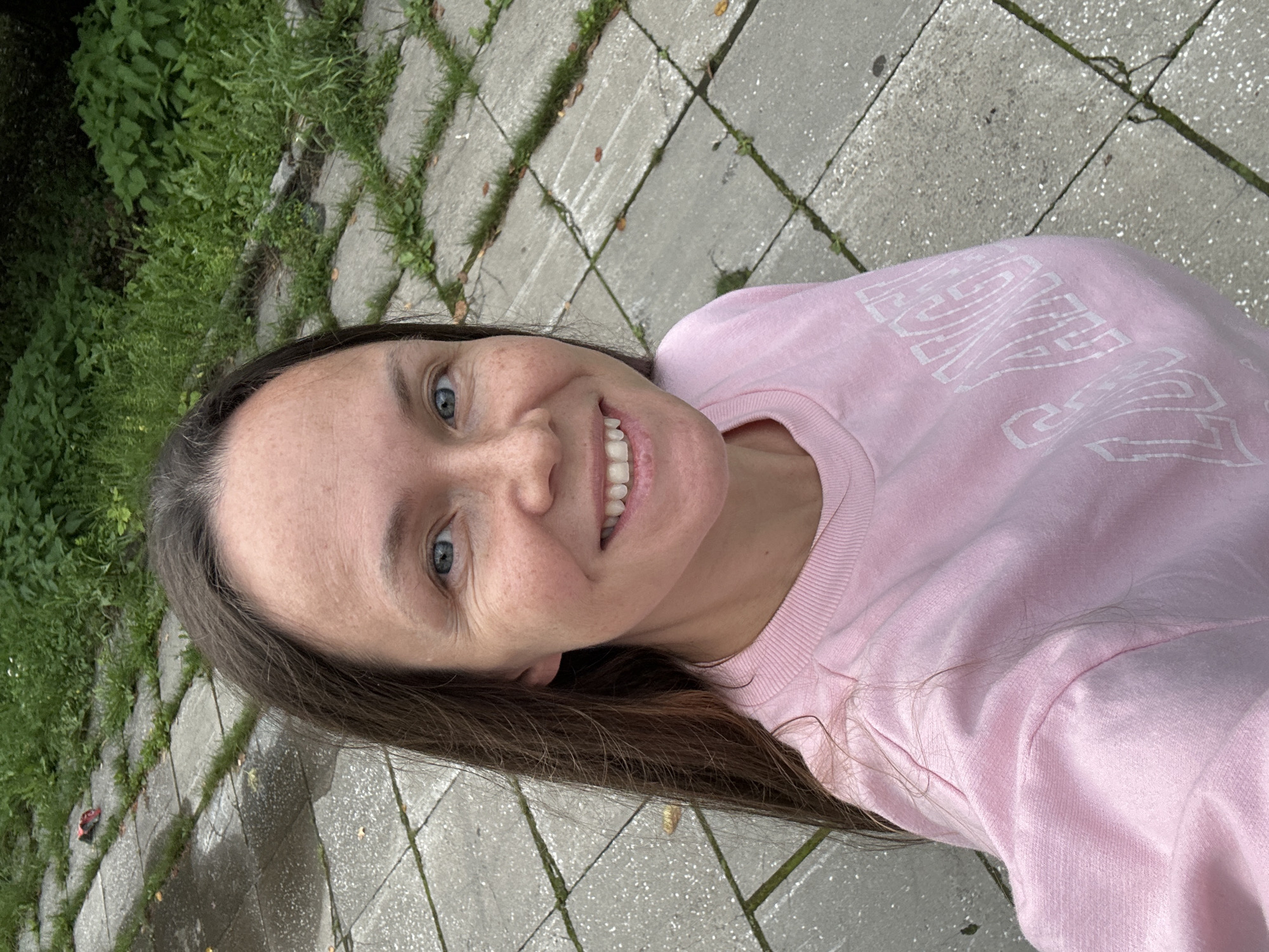 An outdoor, eye-level, selfie-style medium shot shows a woman with fair skin and brown hair smiling and looking at the camera. Her hair is long and falls to her left. She is wearing a light pink crewneck sweatshirt with white lettering that reads "LA ANGEL." The woman's face is close to the camera, and she has light blue eyes and freckles. She has a slight smile, revealing her teeth.
The background consists of a paved area with square, gray concrete slabs, separated by thin strips of grass. Some of the slabs appear wet, suggesting recent rain. Beyond the paving, there is a dense area of green foliage. The overall setting is an outdoor urban environment, likely a park or pedestrian walkway. The lighting is natural, indicating it is daytime. No specific landmarks in Yekaterinburg are visible.