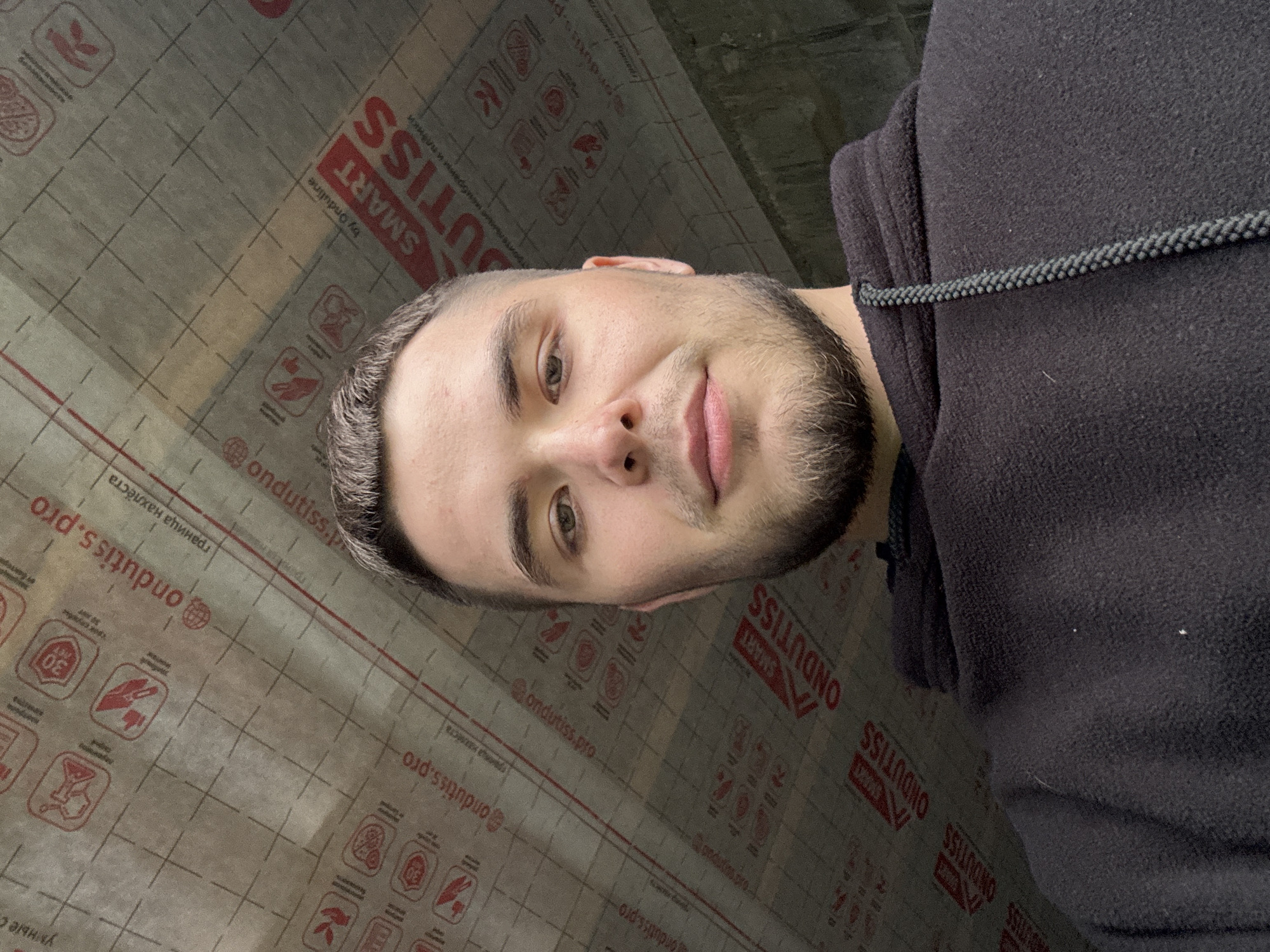 The image is a close-up selfie of a young man in a dark hoodie. He is the primary subject, with his face prominently featured and turned towards the camera. The background consists of a roll of construction material, likely roofing or waterproofing membrane, featuring various logos and text, including "ONDUTISS" and "SMART". The material appears to be a light gray with a grid pattern and red accents, suggesting it's a technical product.
The man has short, dark hair and a beard. His expression is neutral, with his eyes looking directly at the camera. The lighting suggests an indoor setting, possibly a construction site or a storage area, given the background material. There are no clear indicators of the time of day or weather conditions. The overall mood is casual and straightforward. The visible text on the material is in both English and Russian, with brands like "ONDUTISS" and "SMART" repeated, along with terms like "граница нахлеста" (overlap line) and icons indicating usage or safety instructions. The location context provided is Perm, Russia, which aligns with the Russian text seen in the image.