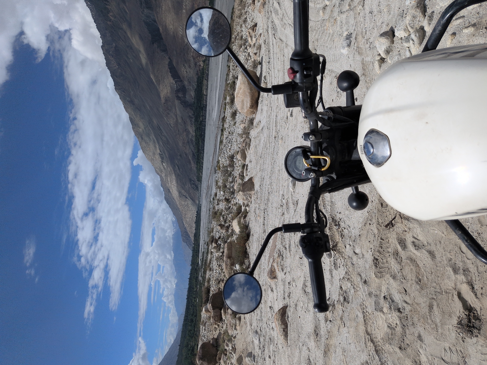 A first-person perspective shot of a white motorcycle is positioned in a rocky, sandy terrain, overlooking a vast, arid valley. The motorcycle's handlebars, mirrors, and part of its fuel tank are visible in the foreground. The left handlebar has a yellow carabiner attached to it. The background reveals a wide riverbed, with sparse vegetation and rocky slopes leading up to imposing, rugged mountains under a bright blue sky dotted with white clouds. The environment appears to be a remote, mountainous landscape, possibly in the region of Sasoma, India, given the user's prompt. No people are present in the image. The scene is static, with no apparent actions taking place. The lighting suggests it is daytime.