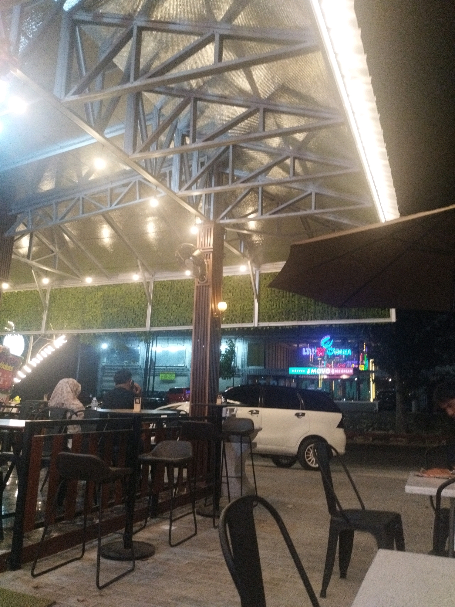 An outdoor seating area is depicted at night, likely a cafe or restaurant, in Sungailiat, Indonesia. The scene is illuminated by artificial lighting, including overhead lights, string lights, and neon signs visible in the background. A metal-framed roof structure with insulating material is visible overhead. In the foreground, several high-top tables and chairs are arranged on a paved surface. To the left, two individuals are seated at a table, one wearing a headscarf. In the mid-ground, a white SUV is parked on the street. In the background, neon signs advertise "LTC CINEMA" and "KOPI MOYO ice cream", indicating commercial establishments. A wall covered in green artificial foliage is also visible. The overall ambiance is that of a casual, nighttime urban setting.