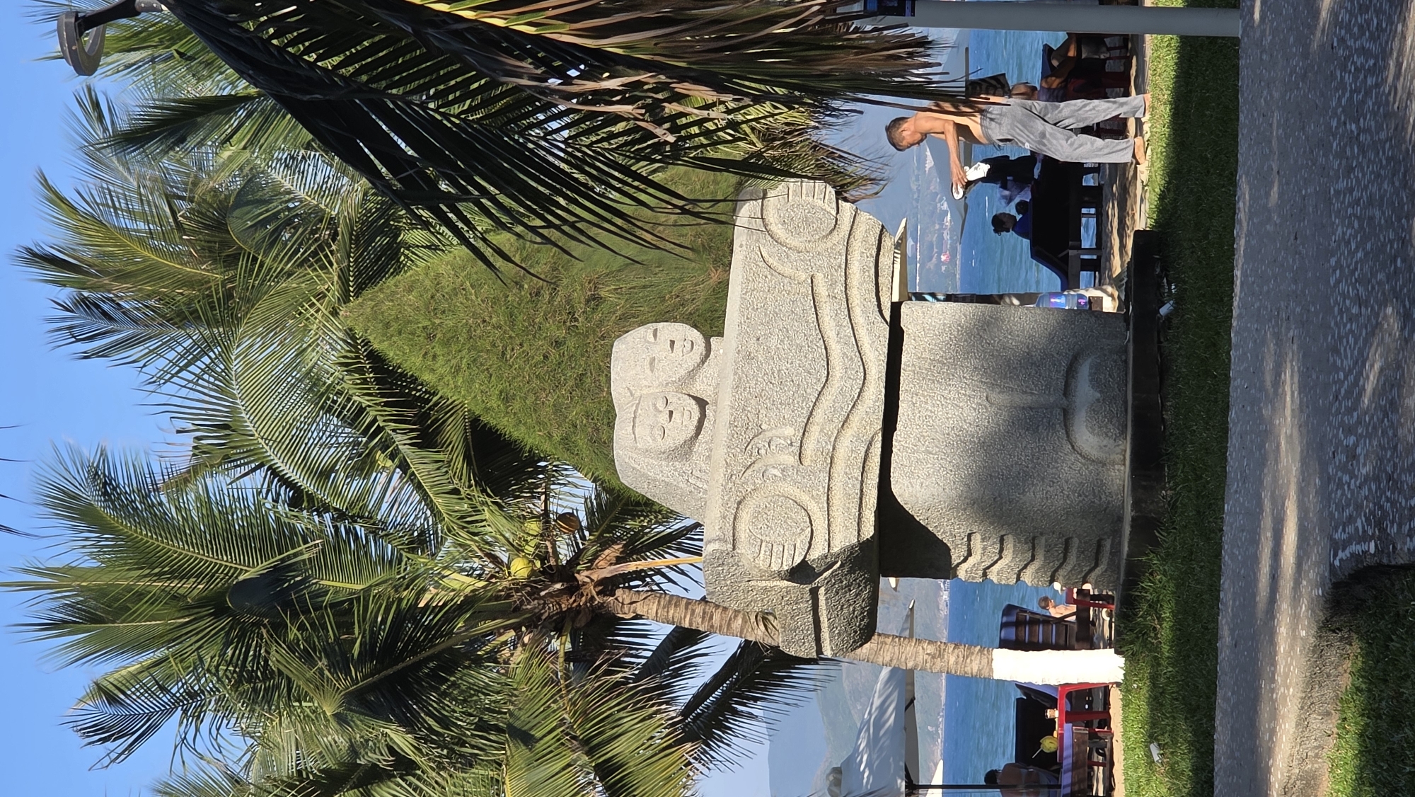 A large stone statue depicting two human heads is the focal point, set against a backdrop of palm trees and the coastline of Nha Trang Ward, Vietnam. The statue is grey, intricately carved with wavy lines and geometric shapes. A man is cleaning the statue with a cloth. The ocean and the city are visible in the background. There is grass and pavement near the statue.