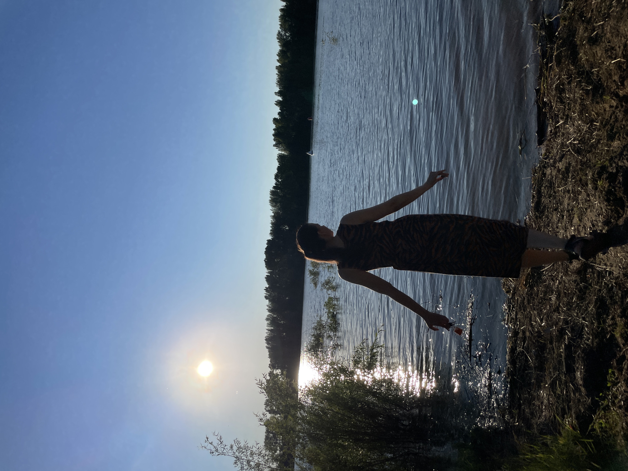 The photograph shows a young woman standing by a body of water, facing away from the camera. She is wearing a sleeveless dress with a tiger-striped pattern and her hair is pulled back in a ponytail. The sun is setting, casting a warm glow on the water and the surrounding landscape. The water appears to be a river or lake, with trees lining the opposite shore. The scene is tranquil and serene, with the woman appearing to be lost in thought or enjoying the moment. The location is the Kama River embankment in Chaikovsky, Russia, known for its picturesque views and recreational opportunities.
