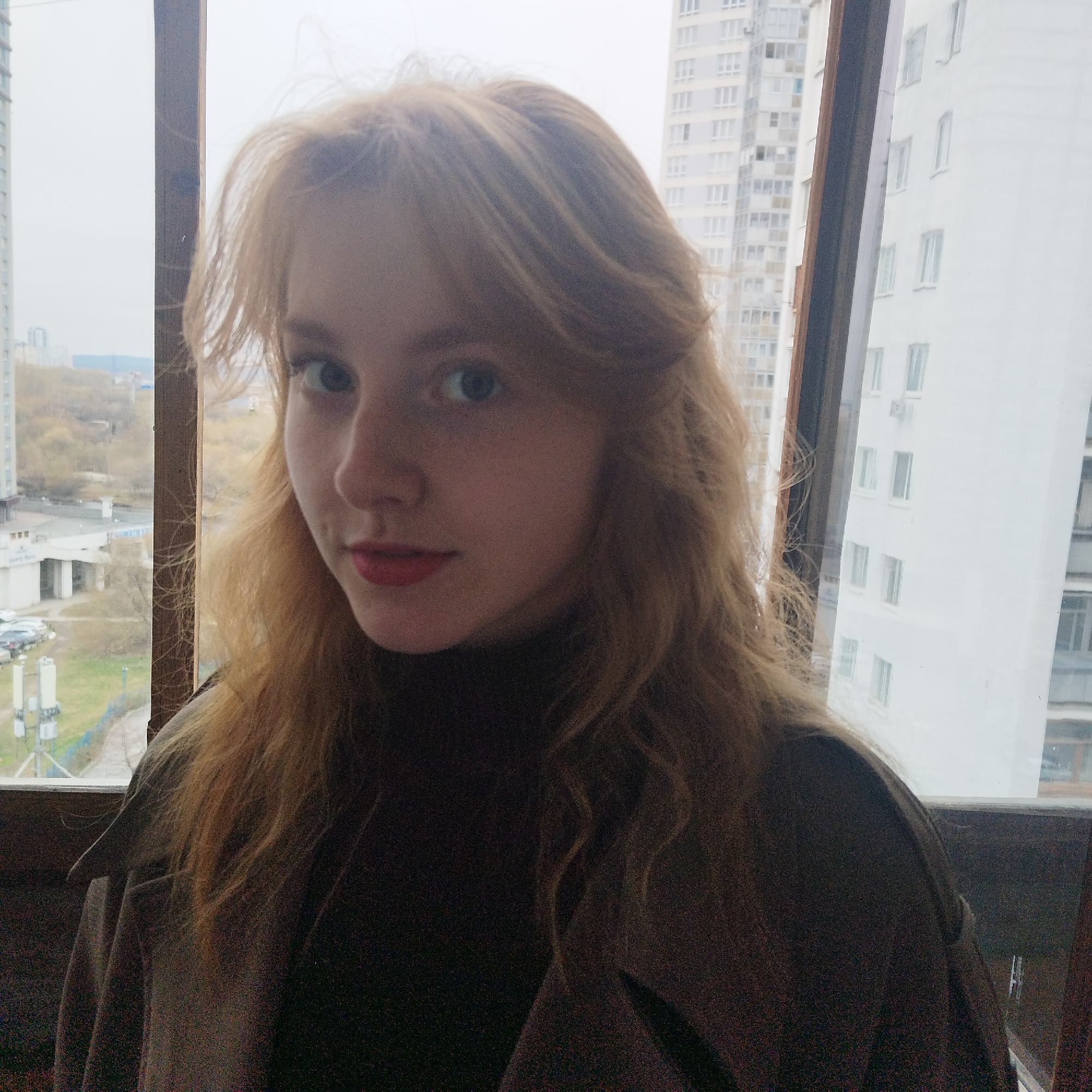 A young woman with long, strawberry blonde hair and light eyes stands in front of a large window. She is wearing a dark brown turtleneck and a similarly colored coat. The background shows a cityscape, typical of urban environments.
Specific clues suggest this photo was taken in Yekaterinburg, Russia. Through the window, one can see several apartment buildings. The architectural style of these buildings is characteristic of post-Soviet construction in Russia. A building with a green roof is visible in the distance; this is a distinctive feature of some areas in Yekaterinburg. Additionally, the overcast sky and general appearance are consistent with the weather conditions in this region.