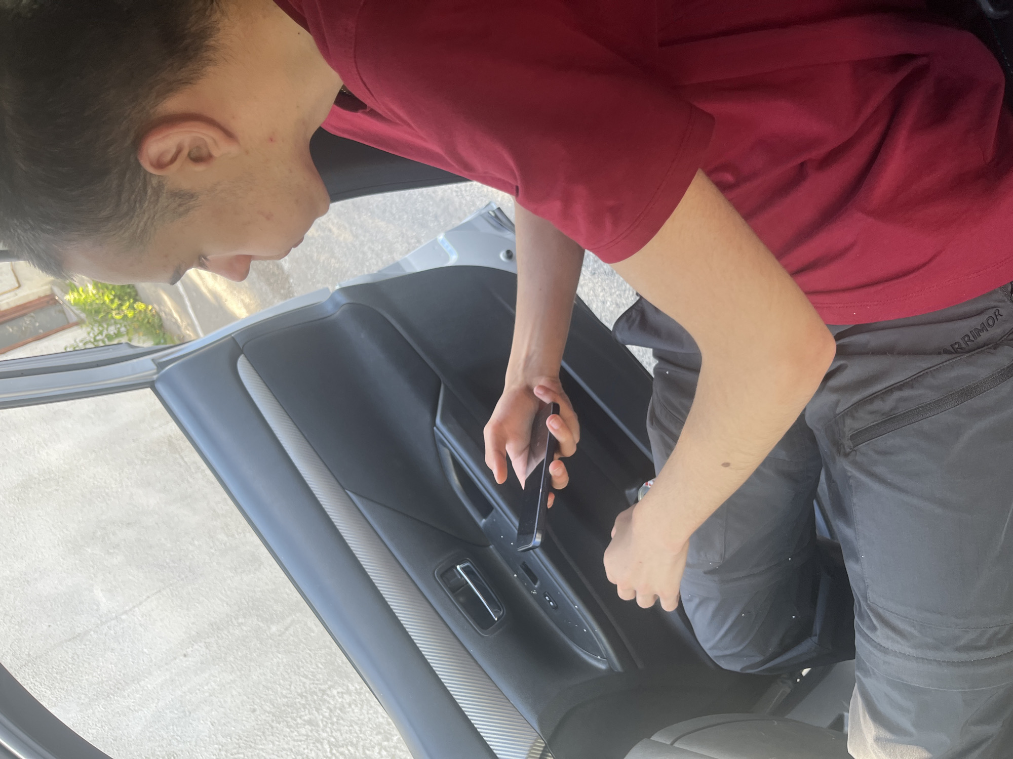 In Stepantsminda, Georgia, a young male with short brown hair, wearing a red t-shirt and grey trousers, is seated inside a vehicle. He is holding a black smartphone in his right hand, appearing to be looking at it. The interior of the car's door panel is visible, featuring a chrome door handle and window controls. The exterior environment outside the car is blurred but suggests an outdoor setting with light-colored pavement and some green foliage.