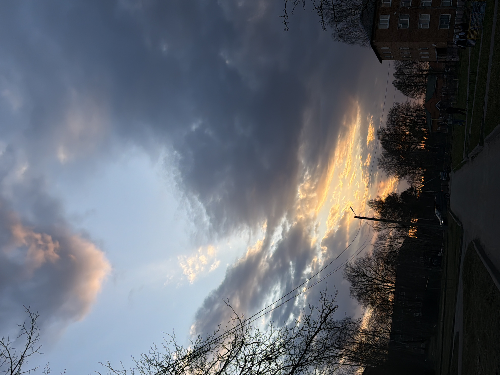 The image shows a sunset sky over Reshetnikovo, Russia. The sky is filled with dramatic clouds, with some illuminated by orange sunlight breaking through. Bare tree branches are visible in the foreground, silhouetted against the sky. A brick building is visible on the right side of the image, along with street lights, and a road.