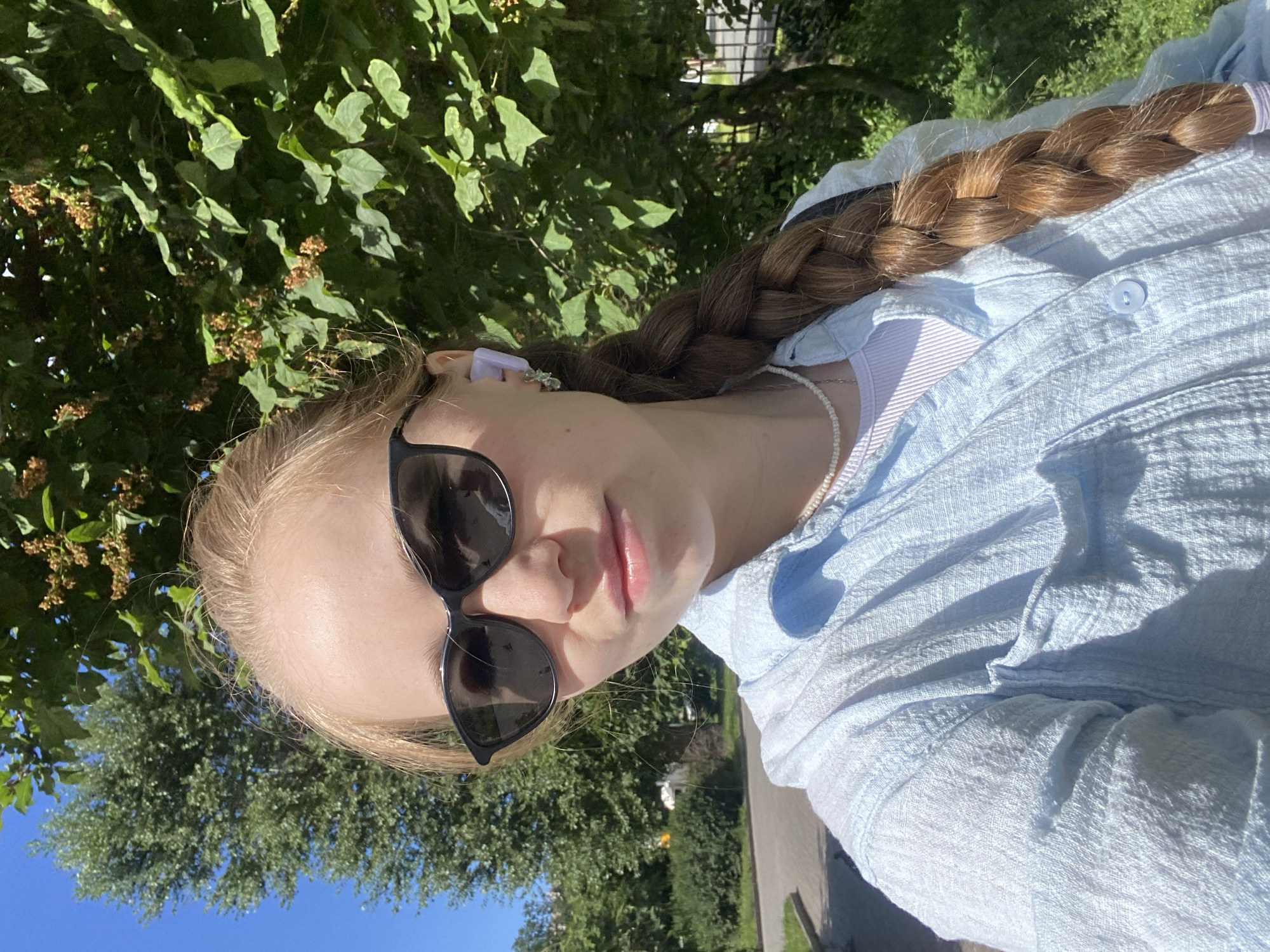 A young woman with blonde hair in a braid is taking a selfie outdoors on a sunny day. She is wearing sunglasses, a light blue button-up shirt over a white ribbed top, and a pearl necklace. She has a purple wireless earbud in her right ear. In the background, there are green trees, a blue sky, and a paved path or road. The overall scene suggests a casual outdoor setting in a park or residential area within Saint Petersburg, Russia.