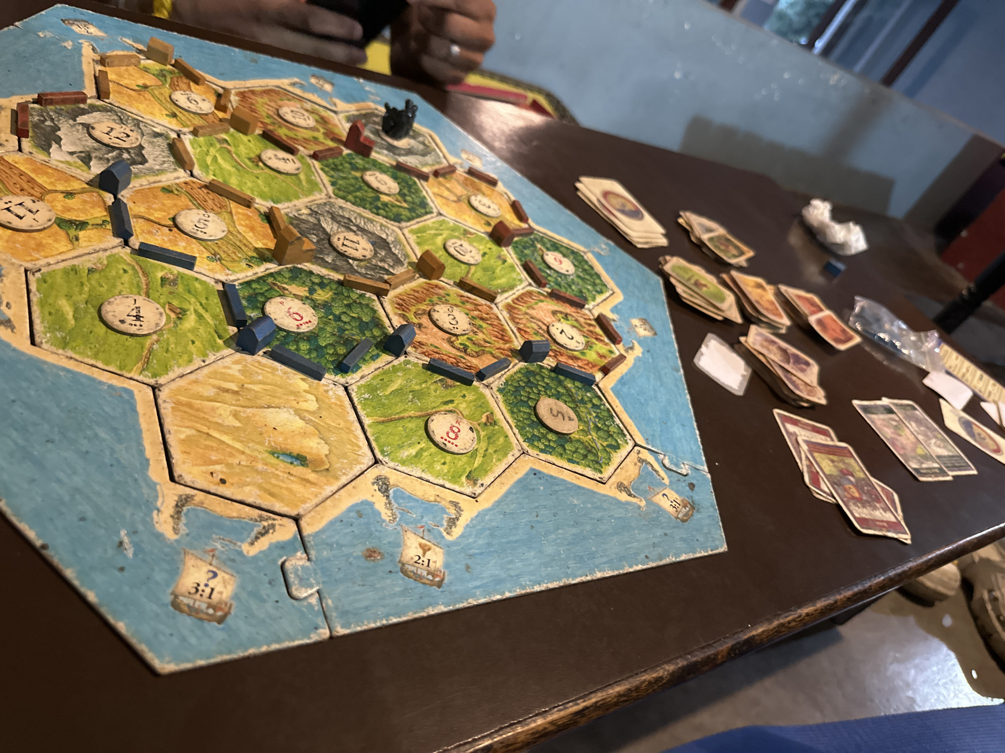 The image shows a close-up, high-angle view of a Catan board game on a dark brown table. The game is set up with hexagonal land tiles representing fields, pastures, mountains, and forests, surrounded by blue sea tiles. Wooden game pieces, including settlements and roads in brown and blue, are placed on the intersections of the hexagonal tiles. Number tokens are visible on some land tiles. Cards are spread out on the table to the right of the game board. A person's hands, one wearing a ring, are partially visible in the upper portion of the image, suggesting they are participating in the game. The environment appears to be indoors, with indirect lighting and a hint of a window or doorway in the background. The overall scene depicts a game in progress.