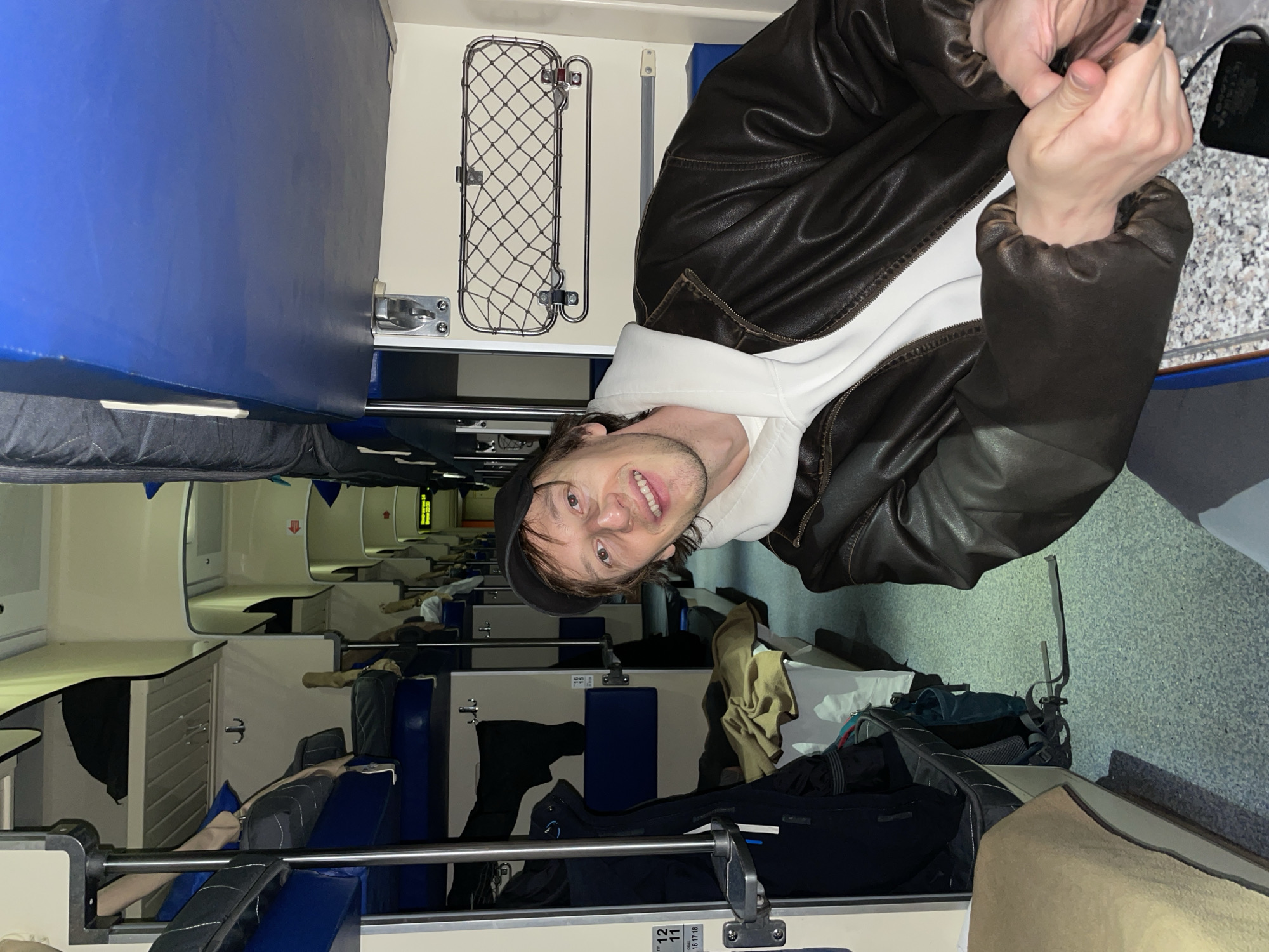 The interior of a train compartment features a man wearing a black cap, white hooded sweatshirt, and brown leather jacket, positioned at the top of the frame, smiling at the camera. A silver device is in his hand. Blue cushioned seats are stacked and covered with various items, including a black backpack, clothing, and other luggage. Overhead, a metal mesh luggage rack is attached to the wall. Further into the train car, rows of compartments can be seen, with blue seating and white walls. The environment appears to be a sleeper train compartment. The location is Unknown, Unknown.