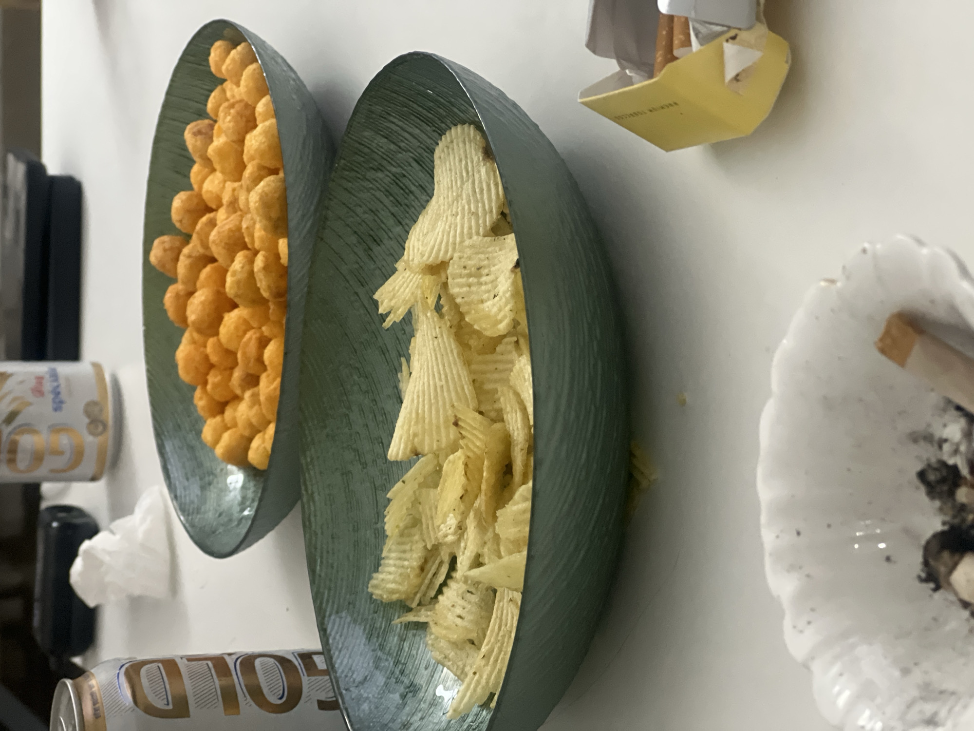 The image displays a casual indoor setting featuring an arrangement of snacks, beverages, and smoking accessories on a white surface, likely a table or countertop. The photograph is oriented vertically, creating the impression that the items are resting on a wall.
In the center of the frame are two long, dark green, textured bowls. The bowl on the left is filled with numerous orange, spherical, puffy snacks, resembling cheese balls or similar puffed corn snacks. The bowl on the right contains light yellow, ridged potato chips, commonly known as crinkle-cut chips.
To the upper right, an open yellow and white cigarette packet is visible, with several cigarettes protruding. The text "PREMIÈRE TOBACCO" is partially discernible on the side of this packet. Below and to the right of the chip bowl, a white, shell-shaped ashtray contains discarded cigarette butts and ash, suggesting recent smoking activity.
On the left side of the image, two aluminum beverage cans are present. One is partially visible at the top left, while the other is more prominently displayed at the bottom left. Both cans feature the word "GOLD" in a white and gold design. A crumpled white tissue lies near the bottom "GOLD" can, and a dark, indistinct object is partially visible behind the left snack bowl.
No people are visible in the scene. The items suggest an informal gathering or a relaxed moment, possibly in the evening. The background is a plain white wall. While the context of Casablanca, Morocco, is provided, the image itself does not contain specific visual cues to indicate this particular location.