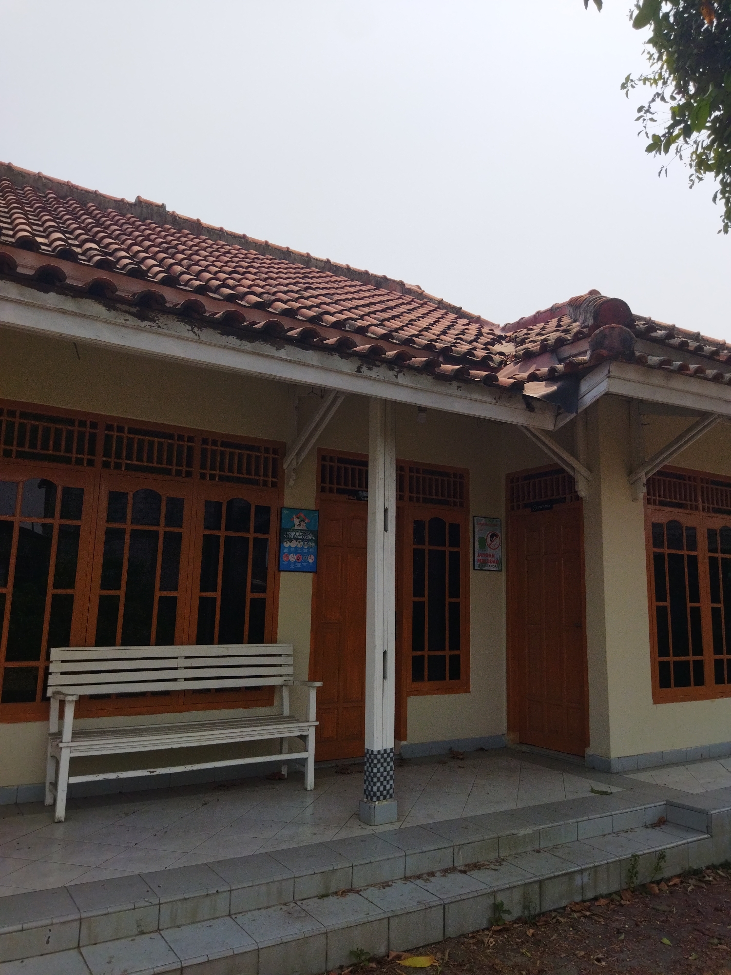 In the city of Suka Sari, Indonesia, a light-colored, single-story building with a red-tiled roof is shown from the exterior. The building has a covered porch area with white wooden benches and large wooden-framed windows. Several wooden doors are visible, some with signs posted on them. The ground in front of the building is paved with square tiles, and there are steps leading down to a dirt area with fallen leaves. The sky is overcast and pale.