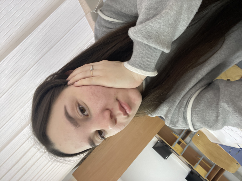 A young female, appearing to be a teenager or young adult, is depicted in a close-up selfie-style photograph. She has long, dark brown hair, a light complexion with visible minor facial blemishes, dark eyebrows, and brown eyes looking directly at the camera with a neutral expression. Her lips are lightly closed. She is wearing a light grey knitted sweater with white trim on the collar and cuffs. Her left hand rests gently on her left cheek, with fingers curled. A ring with a golden band and a central horizontal trapezoidal or diamond-shaped element is visible on the fourth finger of her right hand.
The subject is positioned in what appears to be a classroom, computer lab, or educational setting. In the background, multiple light-colored wooden desks are arranged, each with a black computer monitor. A blue-colored chair is partially visible beneath one of the desks. White vertical window blinds are visible in the upper background, suggesting a window. The walls are light-colored. A piece of paper with writing or diagrams is partially visible on the desk in the foreground, to the lower right.
The scene is situated within an indoor educational facility, such as a school, college, or university classroom/computer lab, in the city of Chaikovsky, Russia. A precise street address or specific name of the institution cannot be determined from the visual evidence.